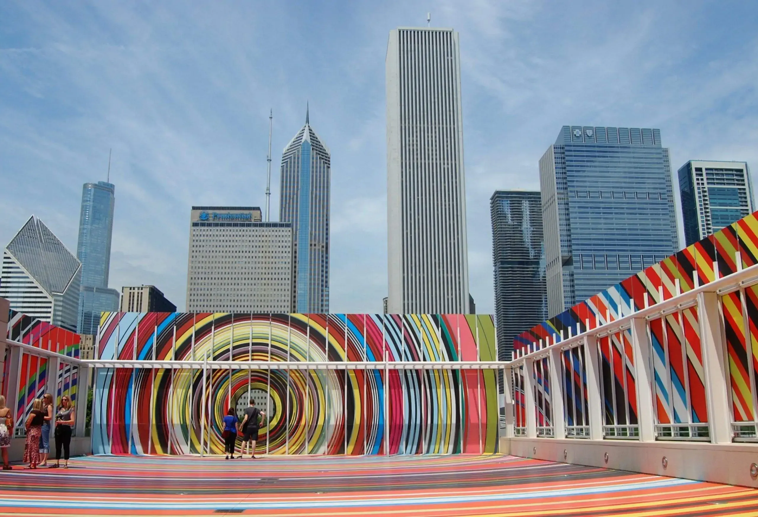 People in an open area with colourful spiral art against the modern towering skyscrapers in the background.