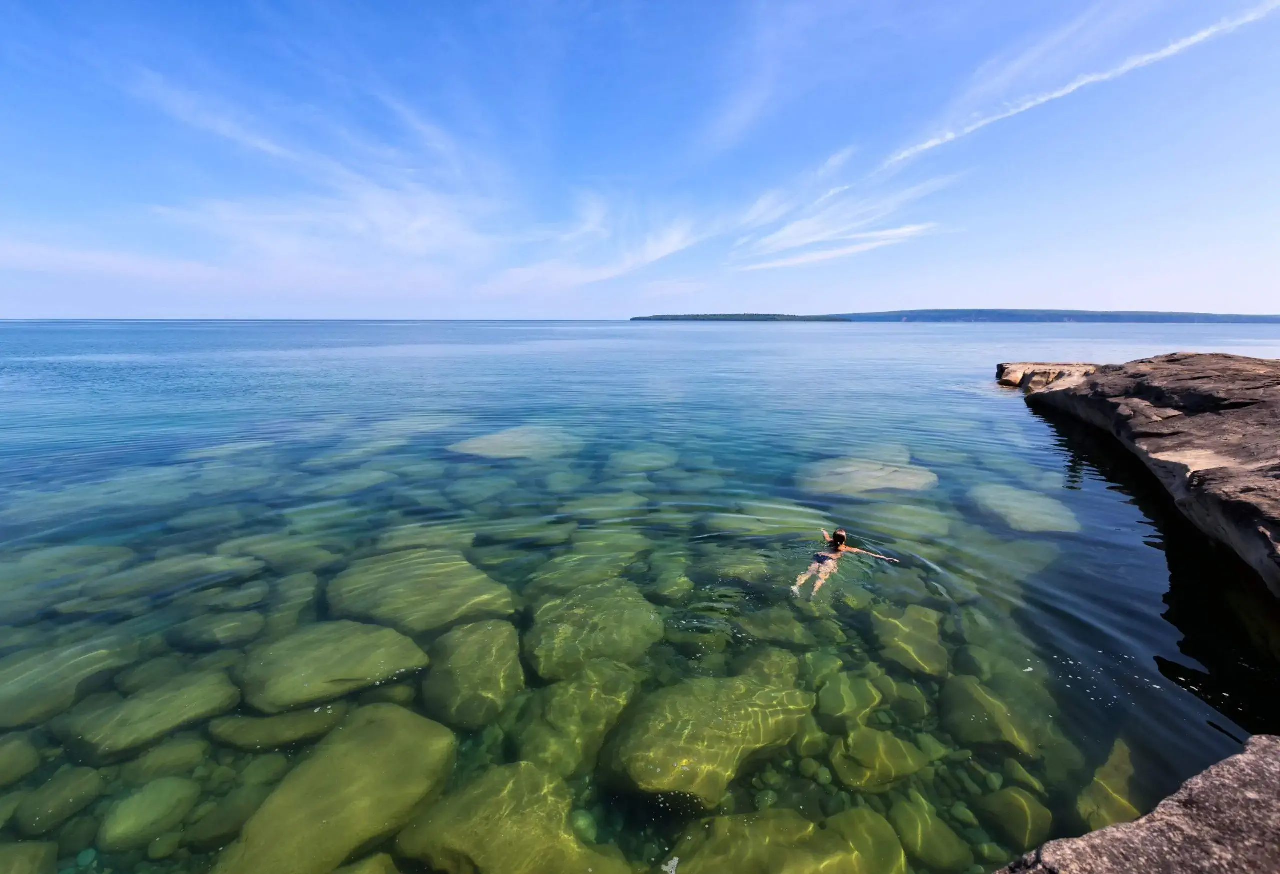 A person swims in a clear lake with large boulders visible beneath the surface.