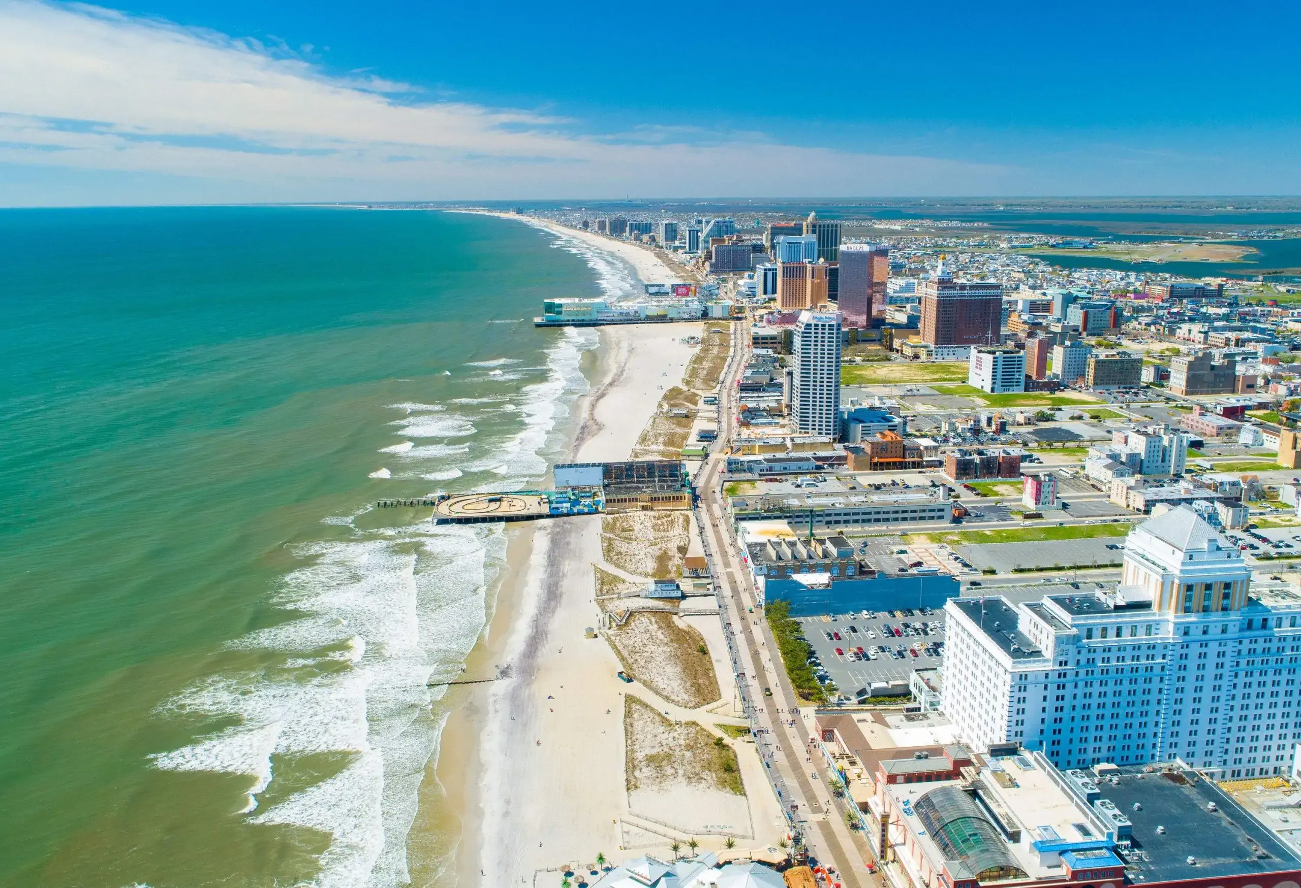 A coastal road cuts across a beach with rough waves and a cluster of waterfront buildings in a city.