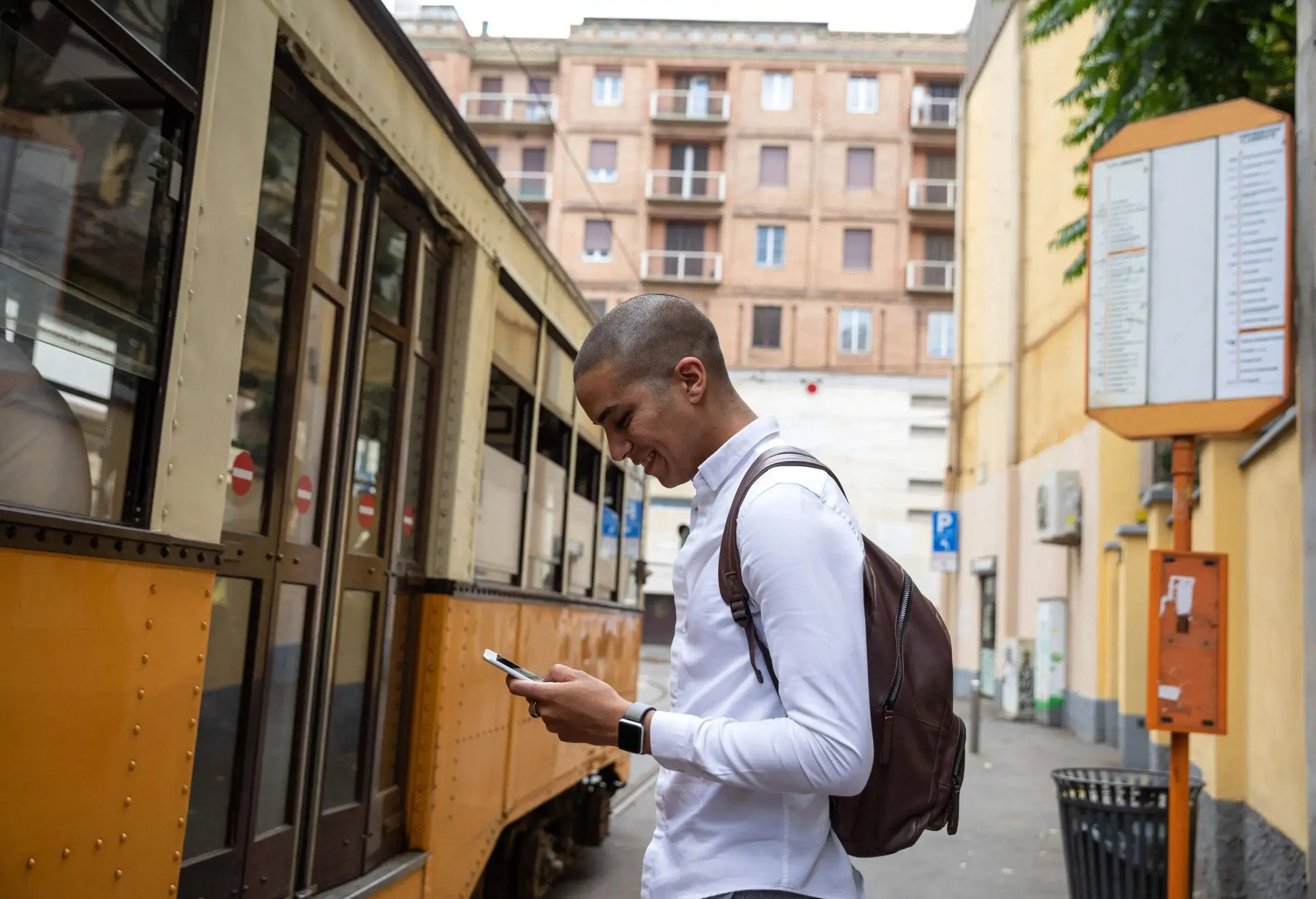 A bald man in a white long-sleeved shirt stands on a bus stop as he smiles at his phone.