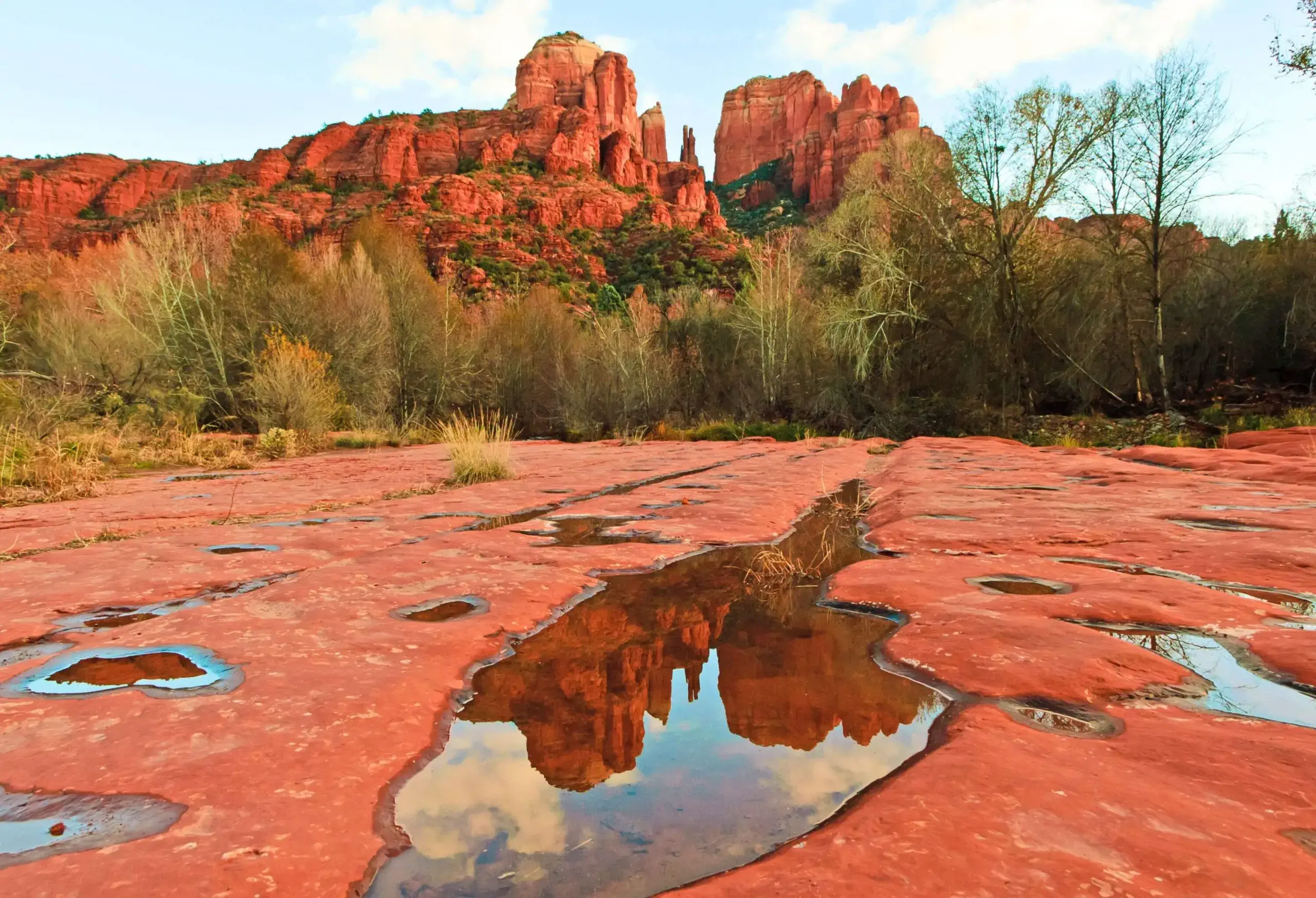 Pools of water on sandstone rocks that reflects the nearby jagged rock formations with steep cliffs.