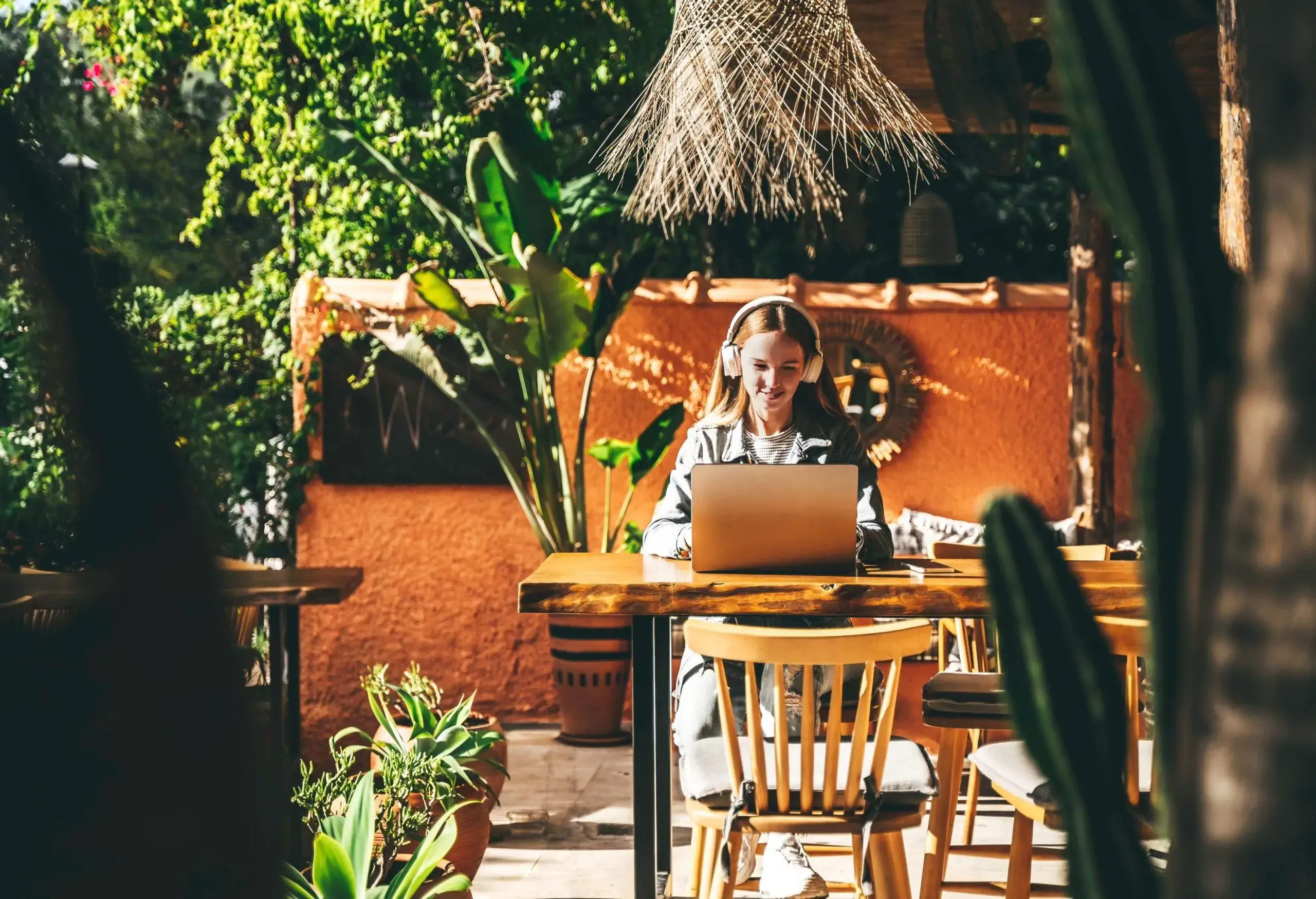 Successful woman is working remotely in a Bali style cafe, organizing the team. She is a digital nomad, happy to spend Winter time in a warm place. Internet and technological communication in terracotta and olive green tones