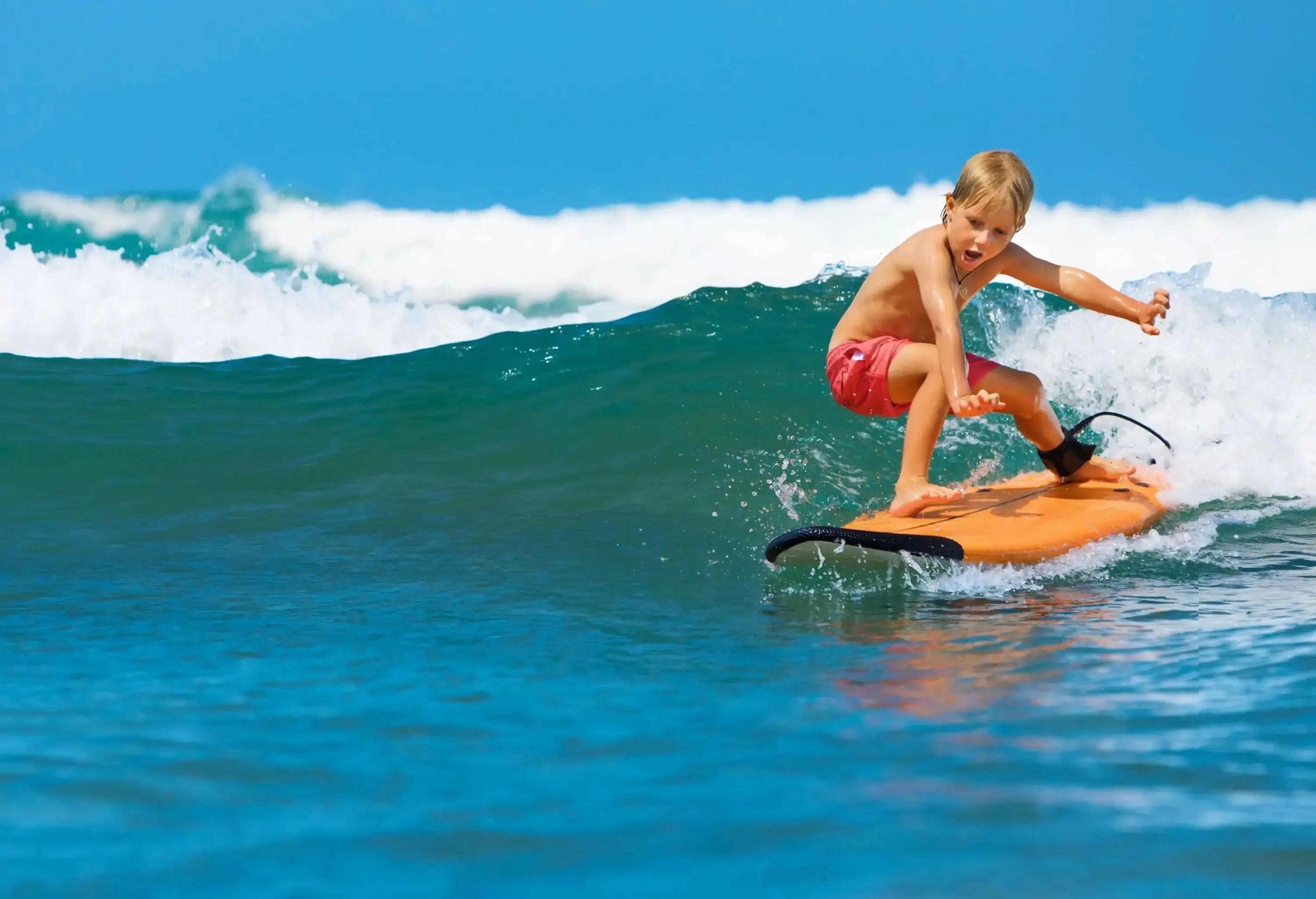 Young blonde kid riding the waves on an orange surfboard.
