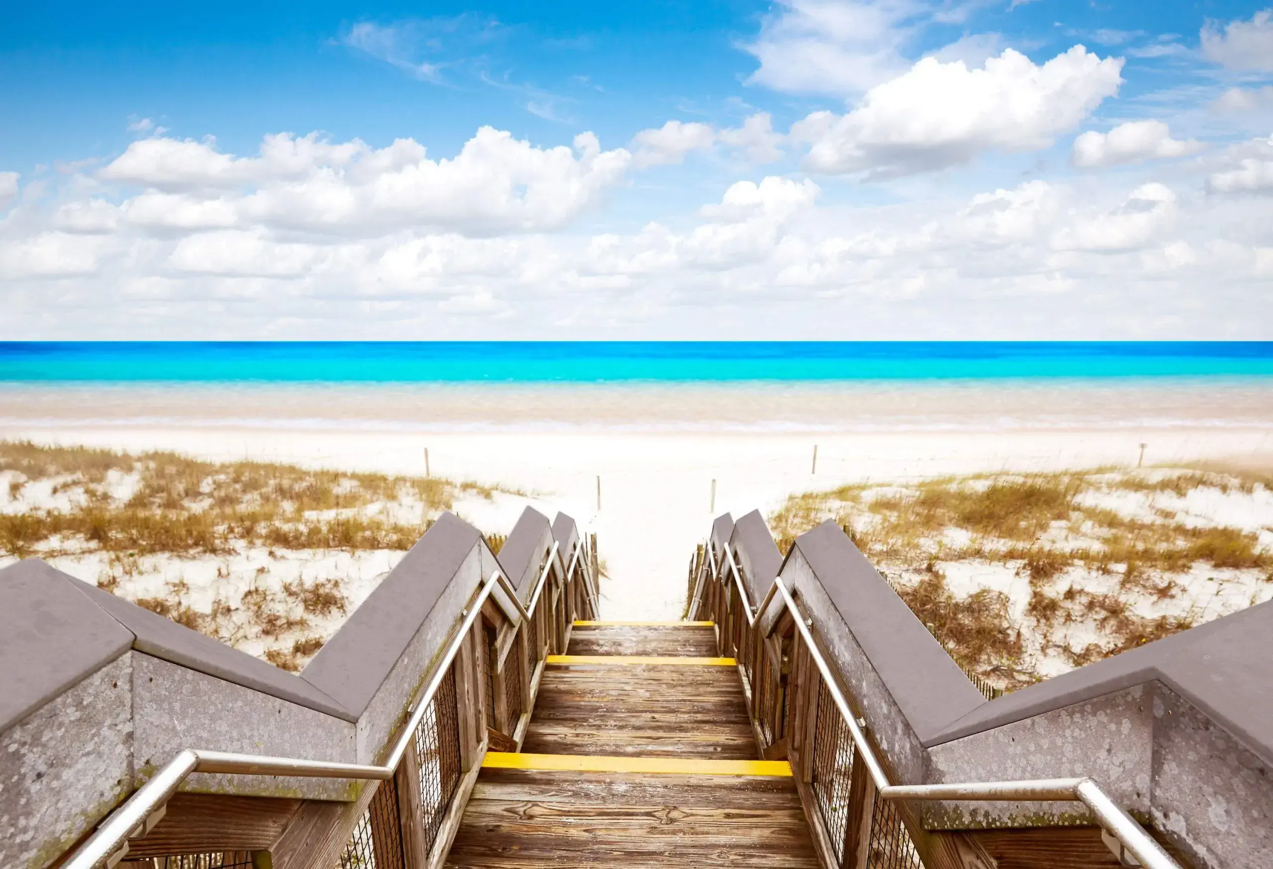 A wooden stair gracefully leads to the shoreline, adorned with scattered grasses, revealing a captivating view of the expansive blue sea beyond.