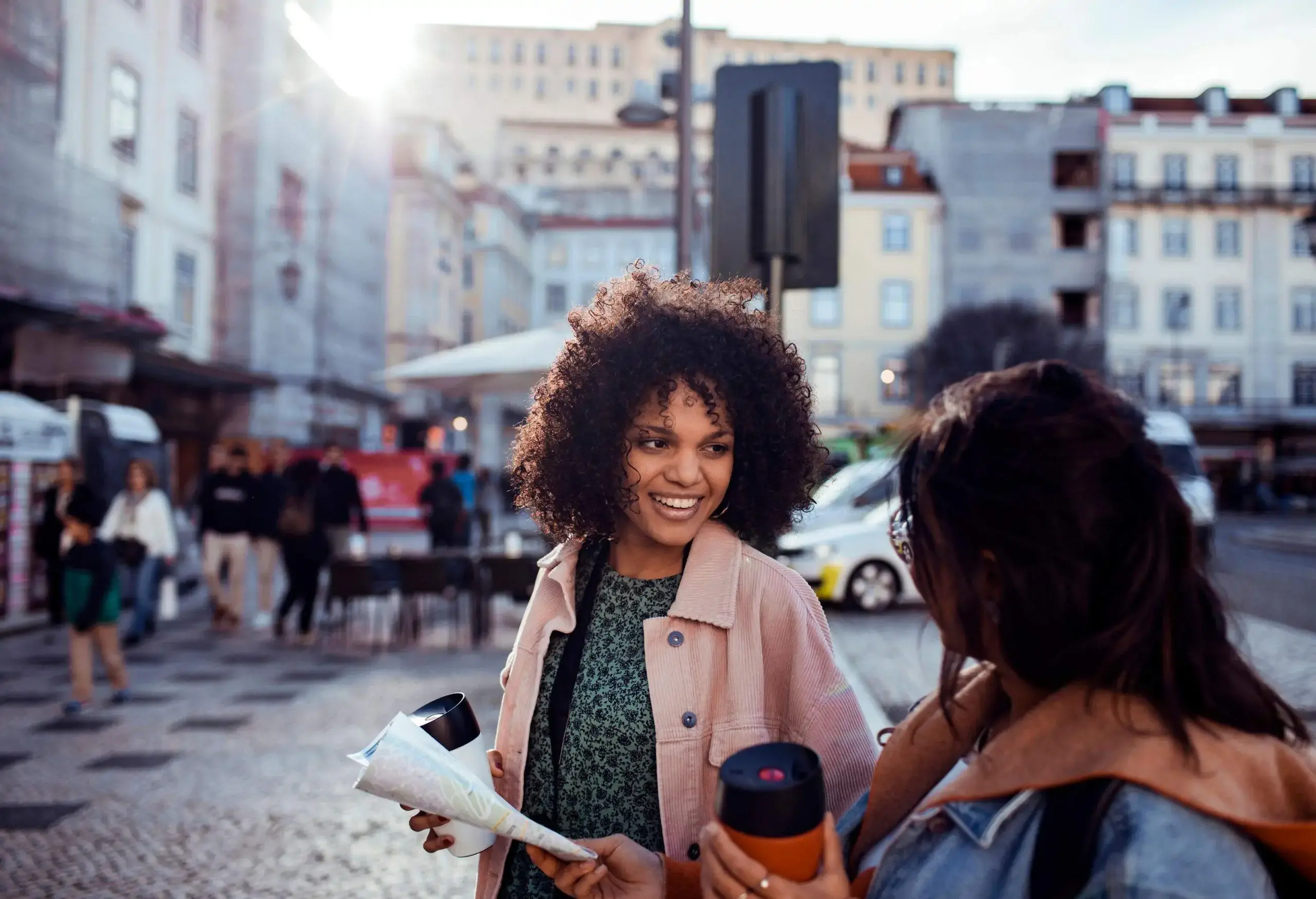 A curly-haired woman smiles at a friend while holding a cup and a map.