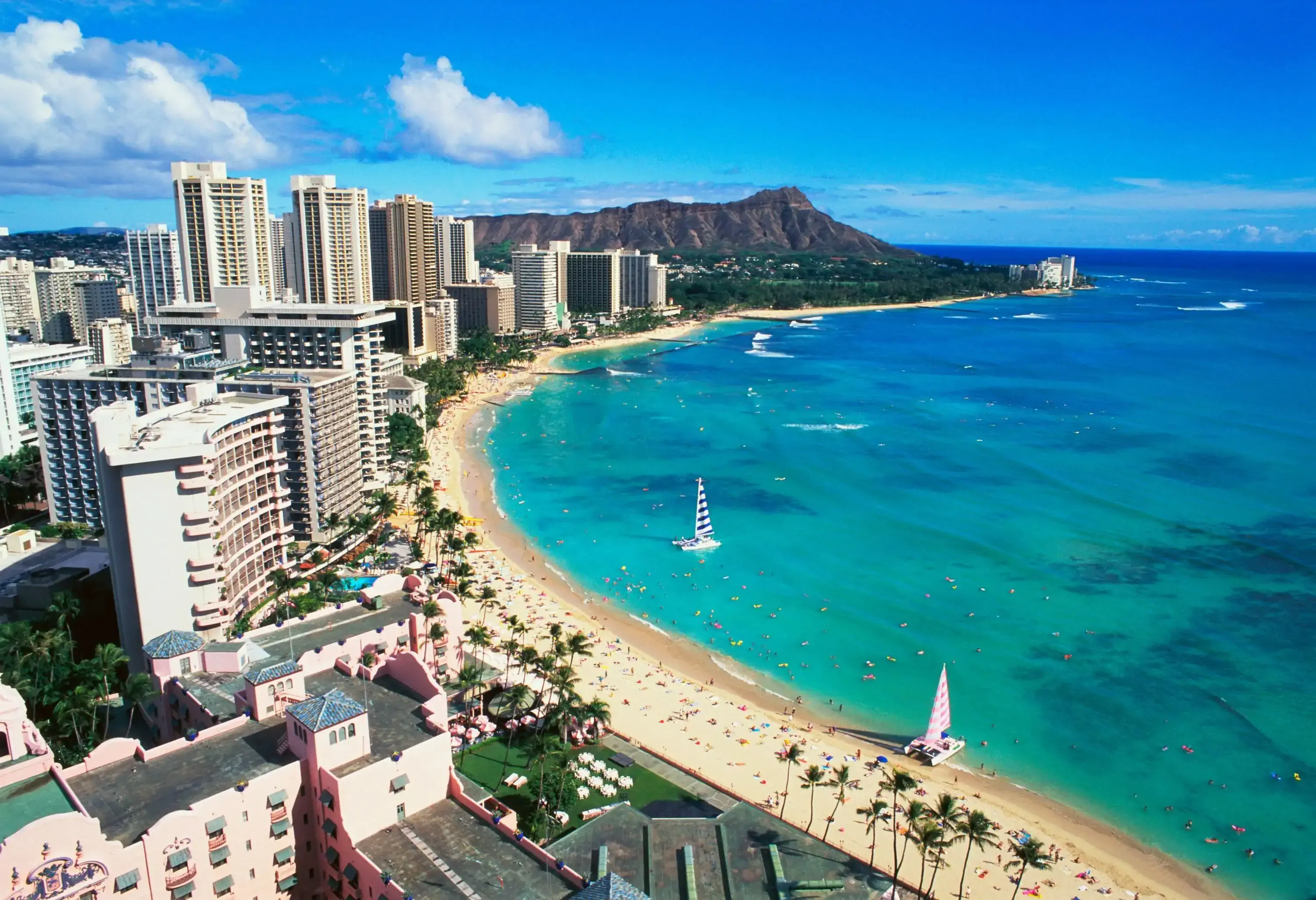 A crescent turquoise beach alongside a cluster of tall compact buildings lined with palm trees.