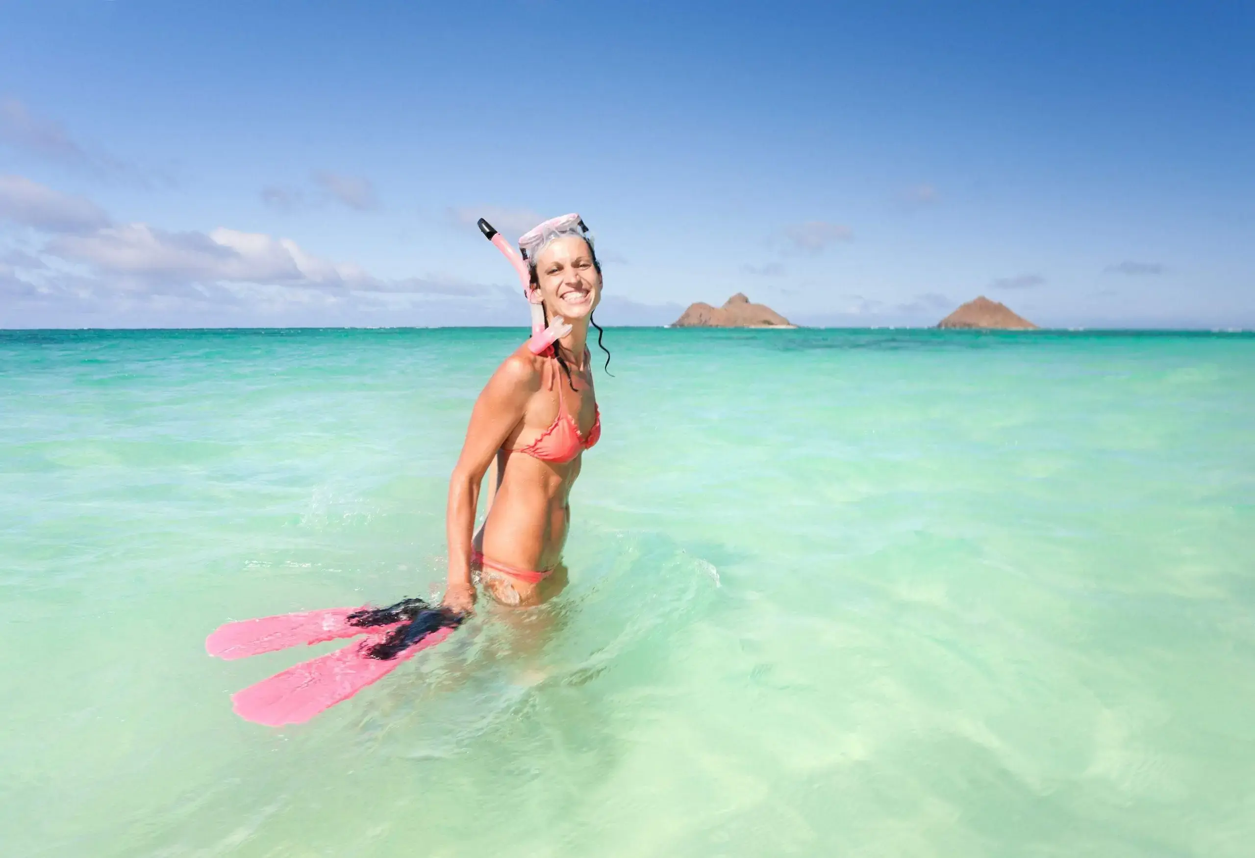 A woman in a bikini with her snorkel gear stands on the sea's shallow waters with two protruding rock mountains in the background.