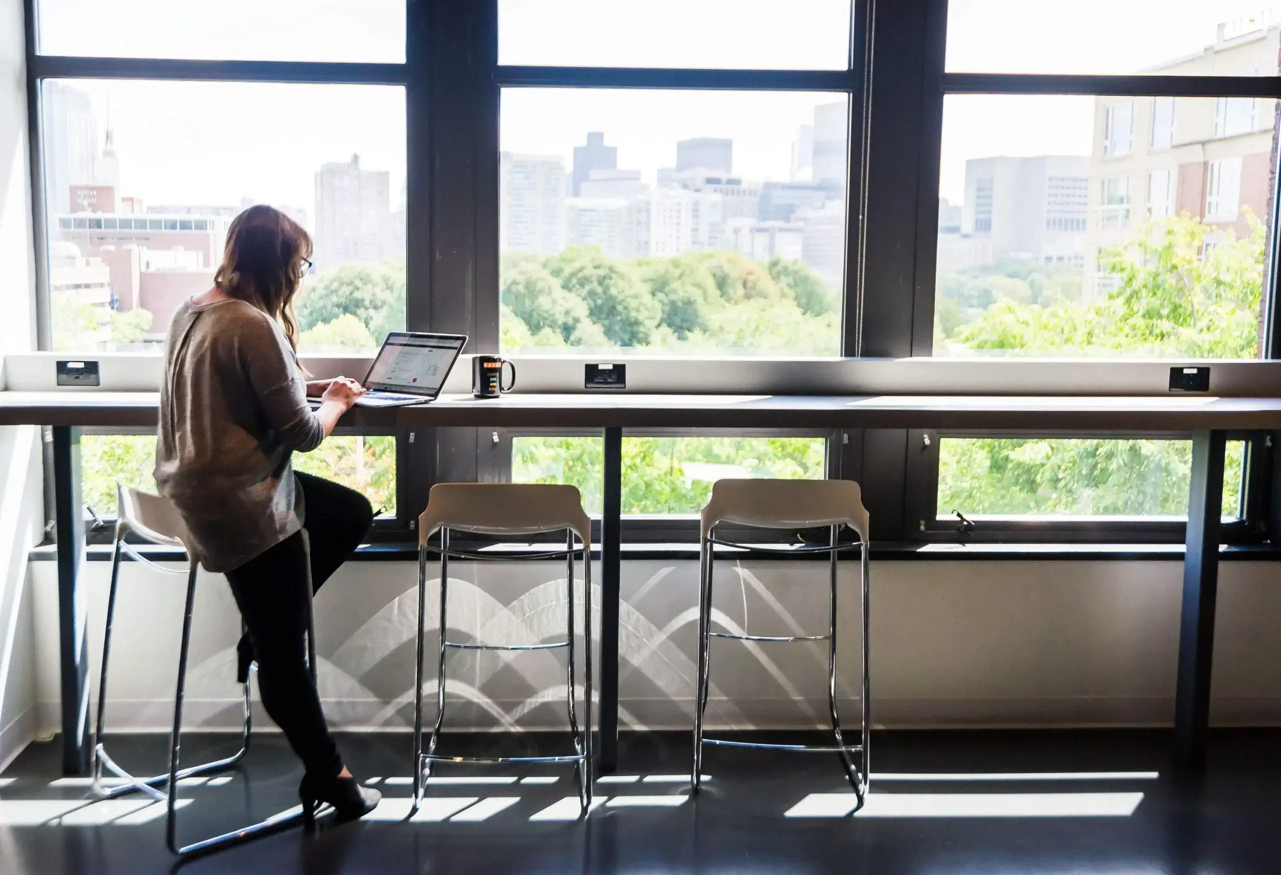 An employee working on her laptop, seated comfortably on a high chair at a high table in the well-lit and spacious office pantry.