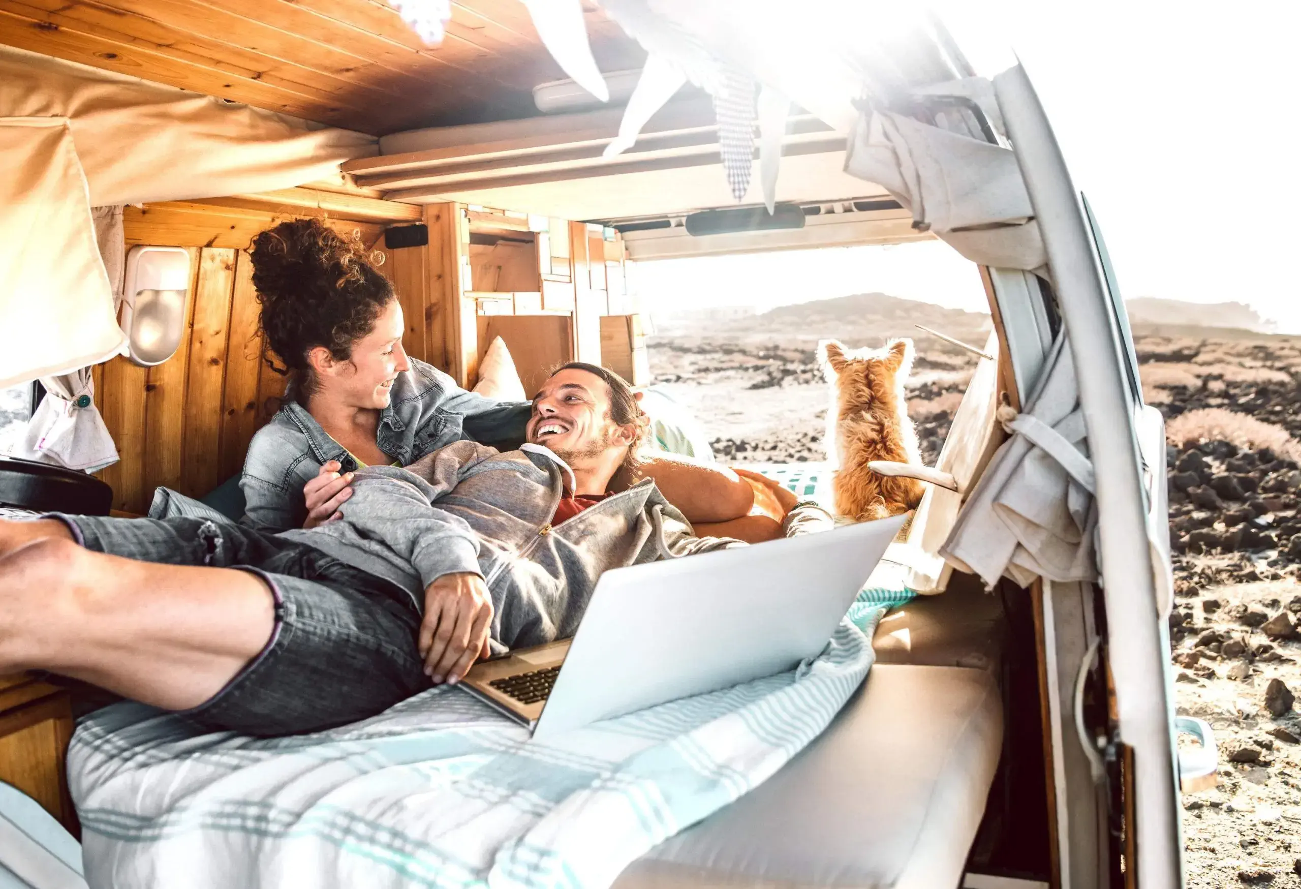 Couple grinning at each other as they lie on a bed inside the van with a dog gazing out the window.