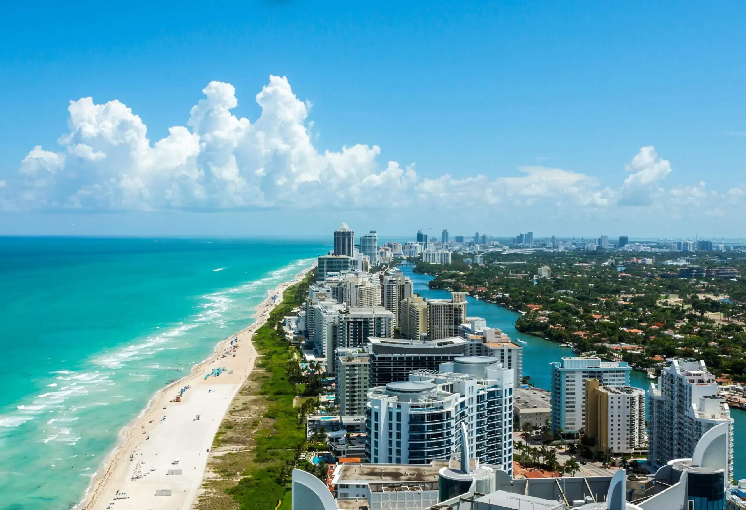 Compact tall buildings form a long line between a tranquil river and the turquoise sea on a bright fine day.
