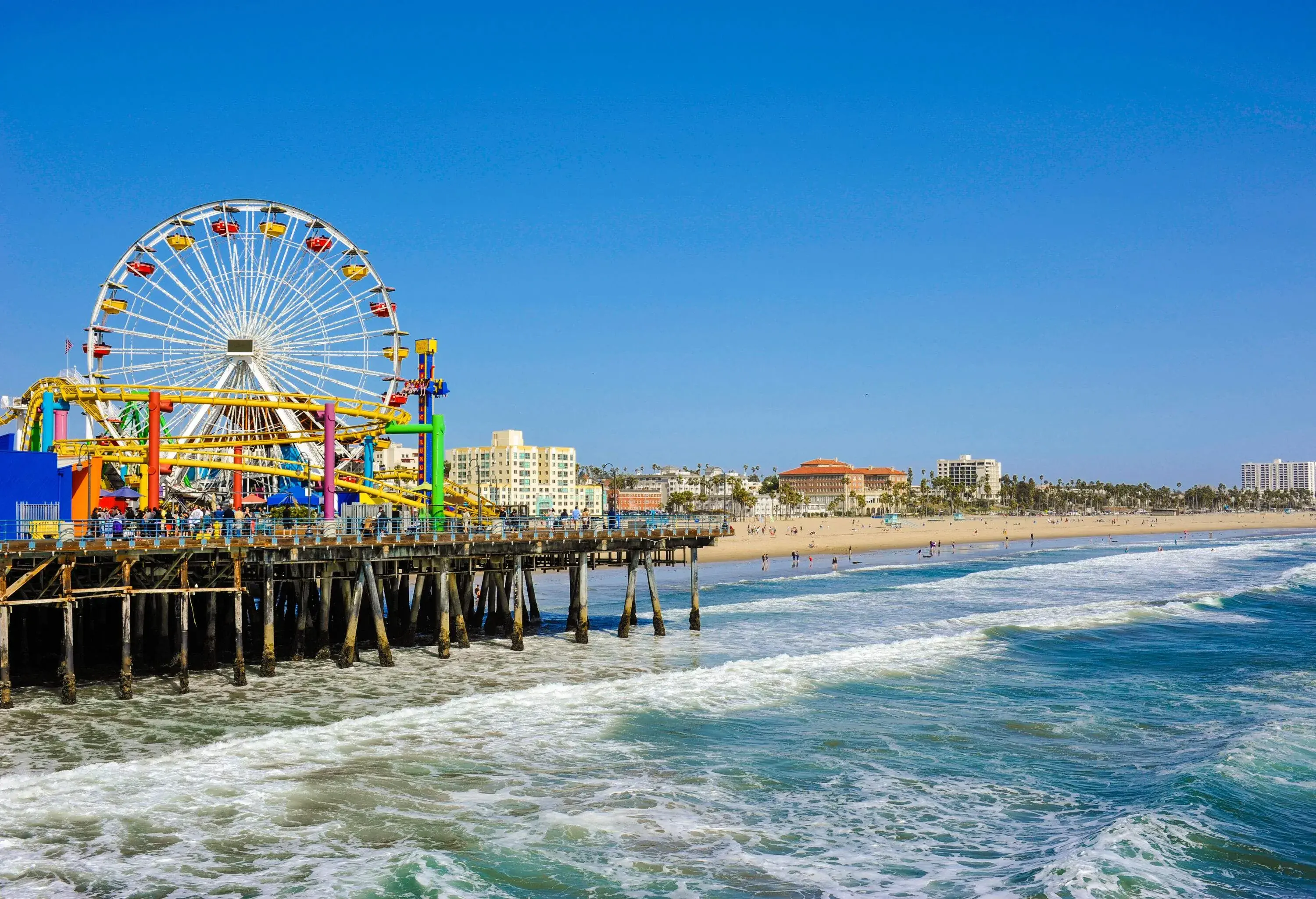 A pier with a colourful amusement park on the shore of a beach along an urban cityscape.