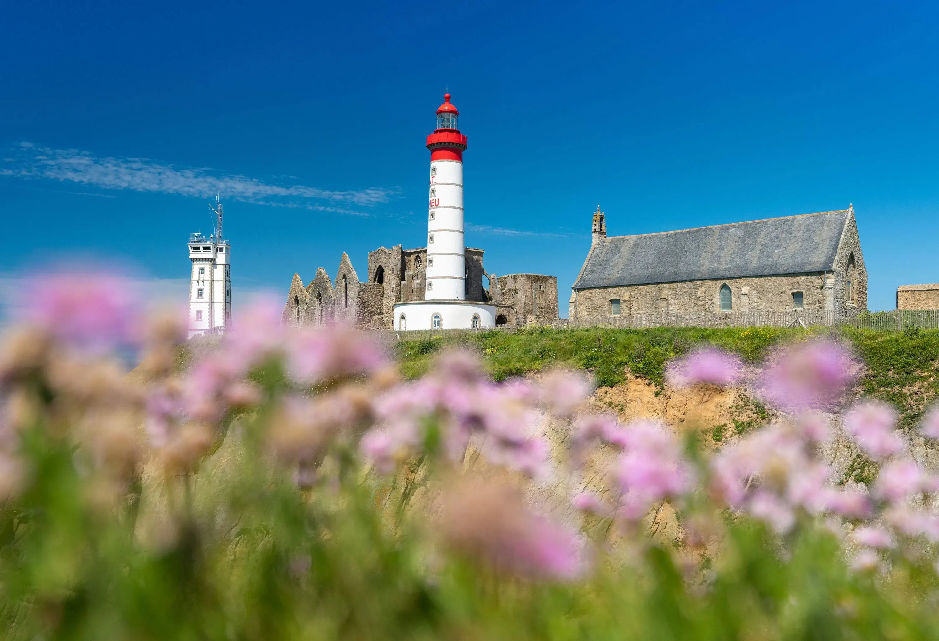 A lighthouse beside the ruins of an abbey and a signal station in the background over a lush green land.