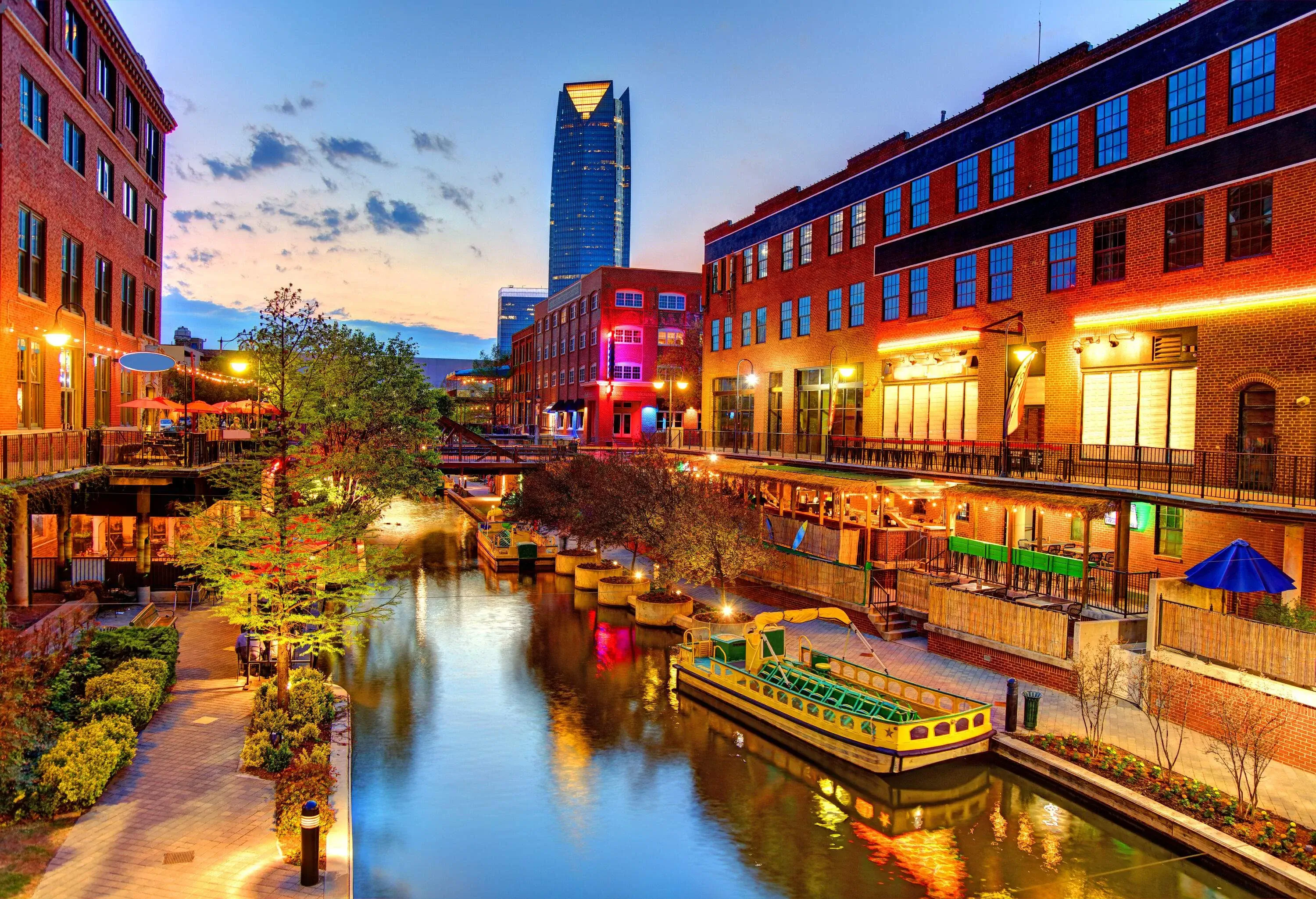 A night-time view of a calm water canal with an anchored tourist boat along a row of trees and commercial establishments.