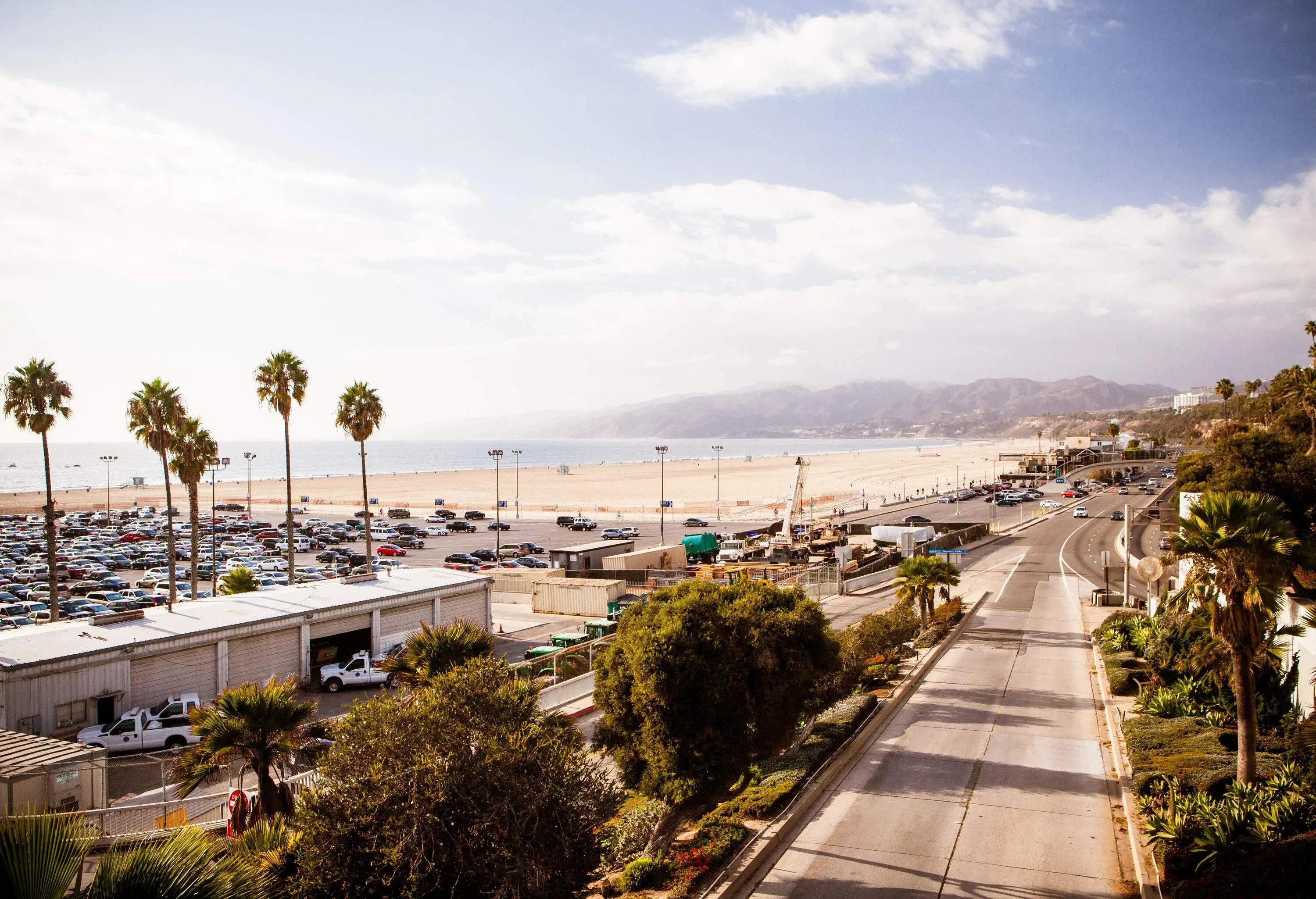 A busy highway along a spacious sandy beach next to a parking lot with an expansive ocean view.