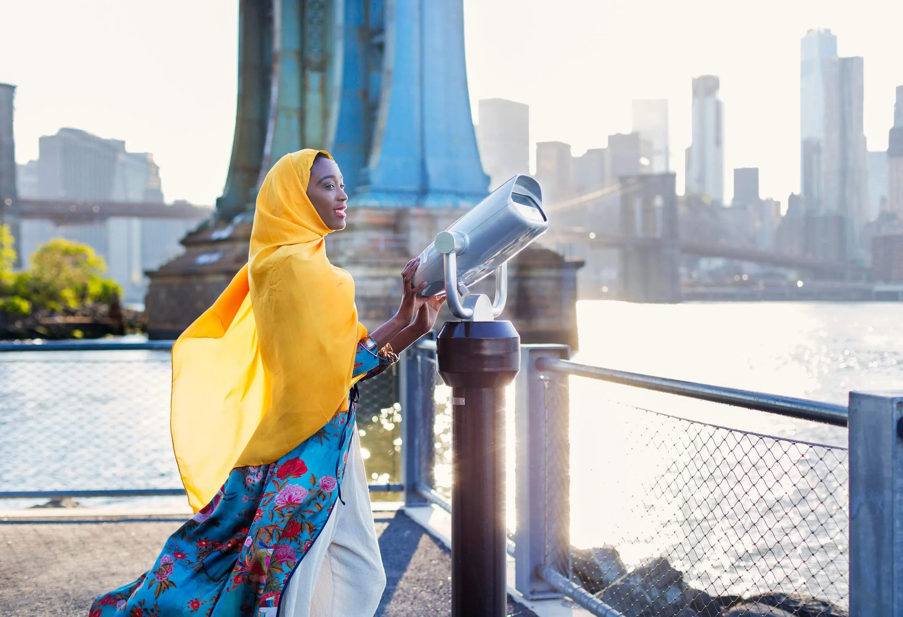 A dark-skinned female tourist wearing a long yellow scarf over her head standing beside a massive telescope in a promenade.