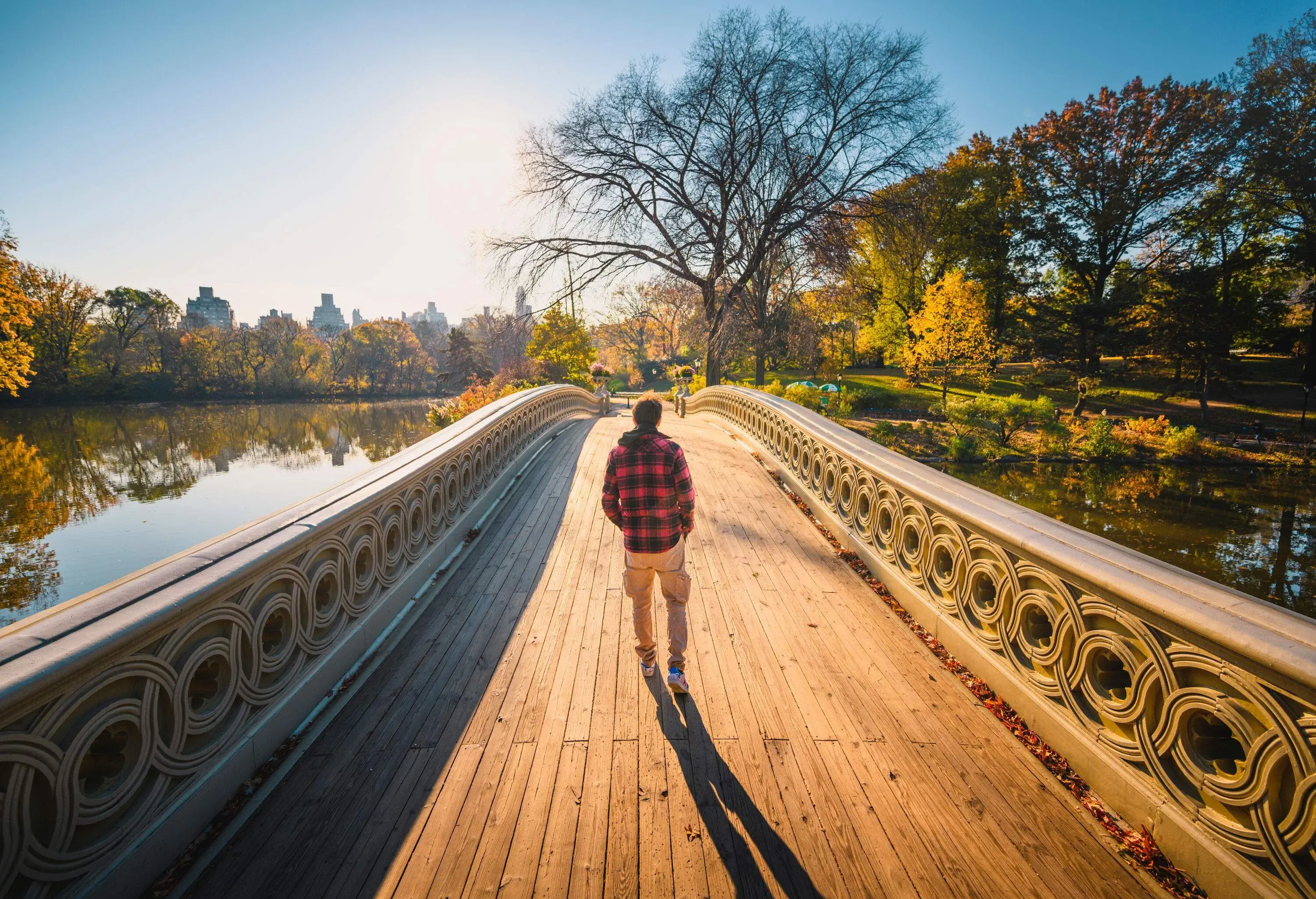 A man in a chequered shirt crosses a footbridge across a calm lake surrounded by vibrant fall foliage.
