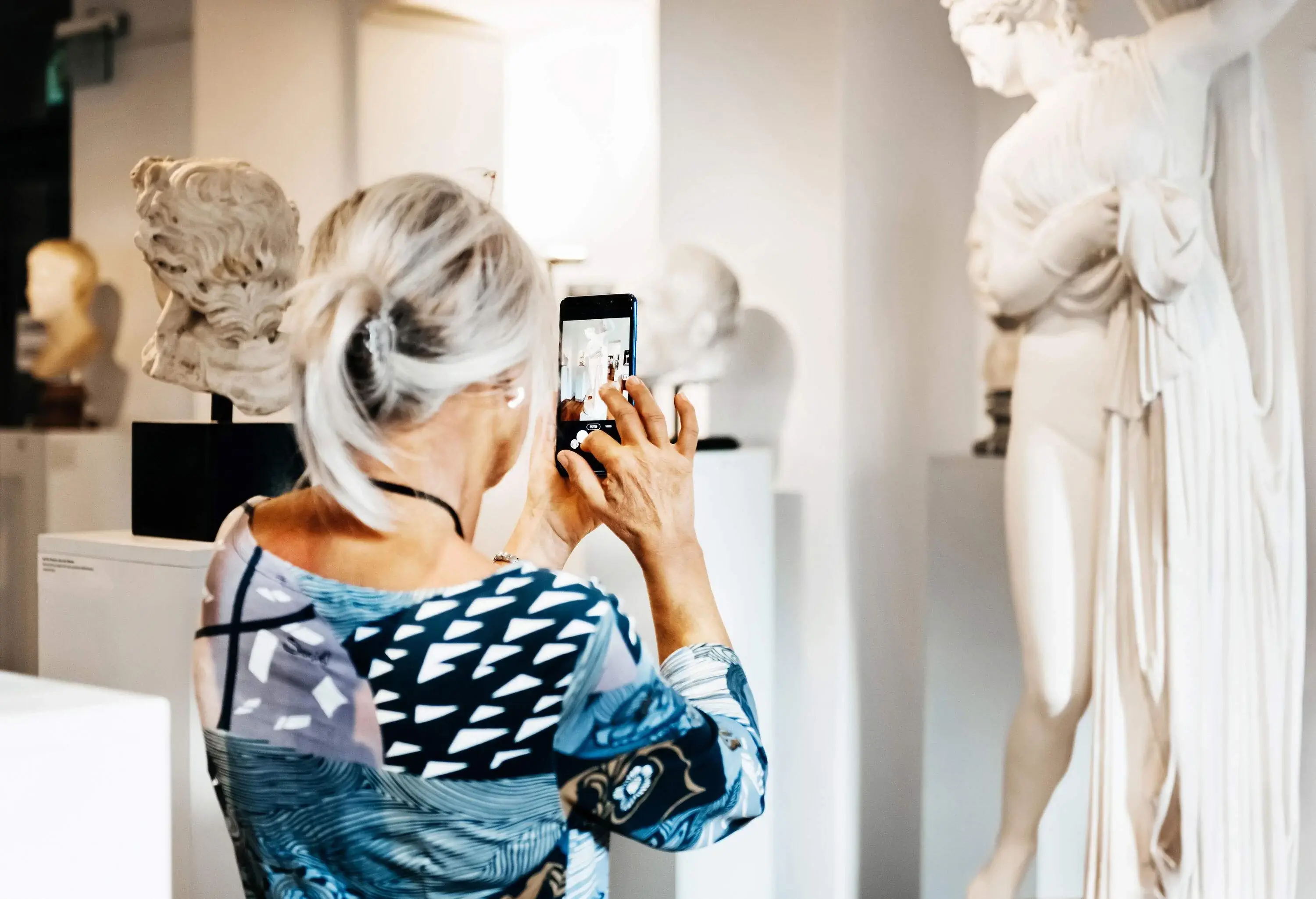 A mature woman expertly captures the beauty of a classical sculpture with her smartphone.