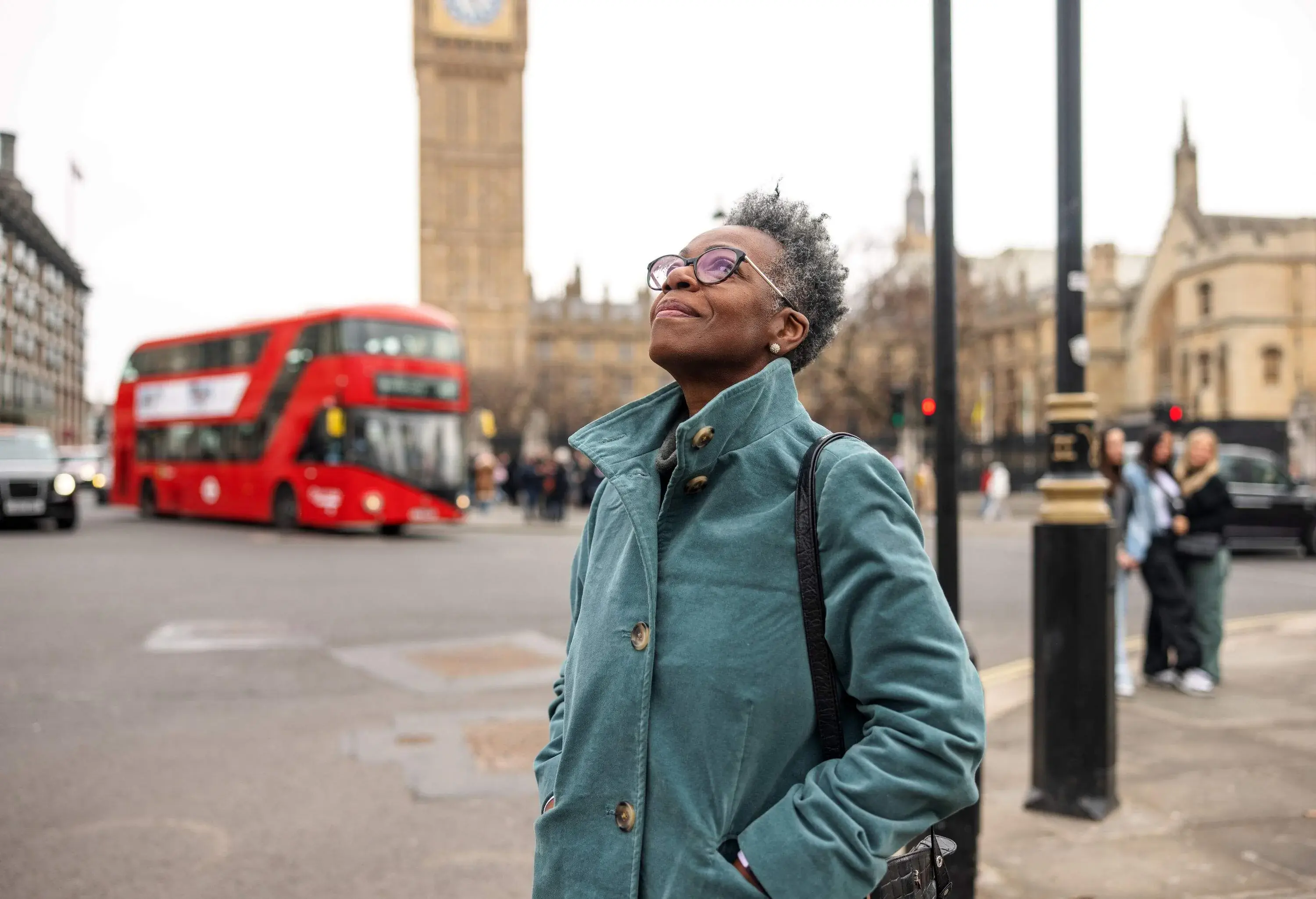 A senior adult black female tourist admiring the architecture in London. She is traveling alone and walking around the City of Westminster on a gloomy winter day. The woman is standing on the sidewalk and behind her, there is the famous Big Ben. She is looking up.