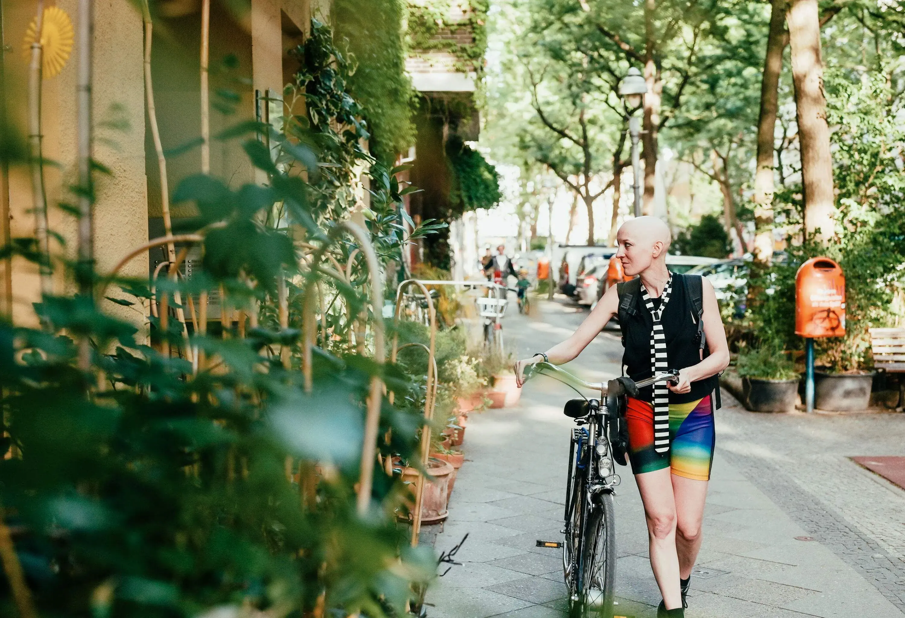 A bald individual with a bicycle in a street lined with buildings adorned with green plants and tall green trees.