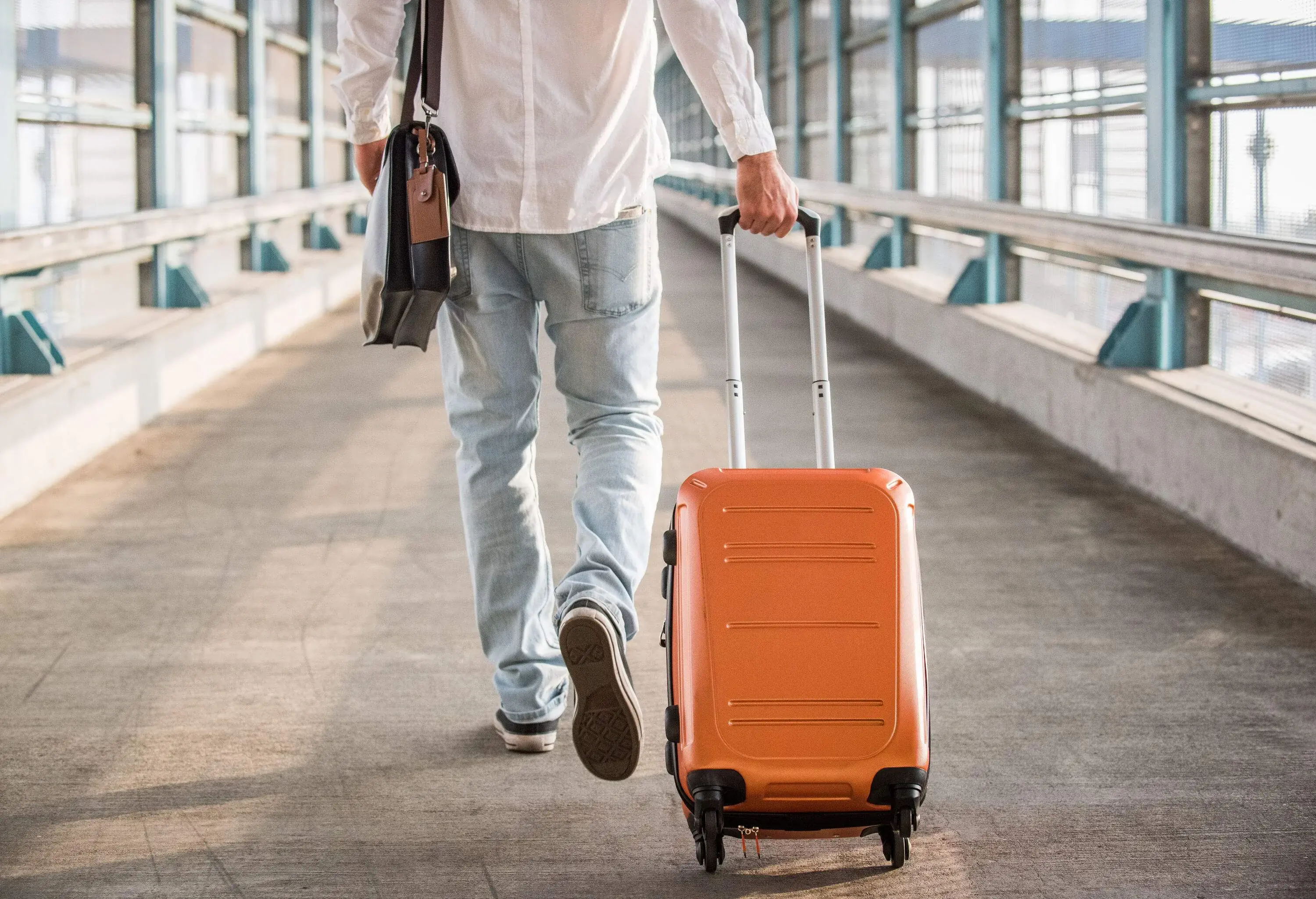 A man wearing casual attire, carrying a backpack and dragging a suitcase, walks along a walkway leading to a railway station.