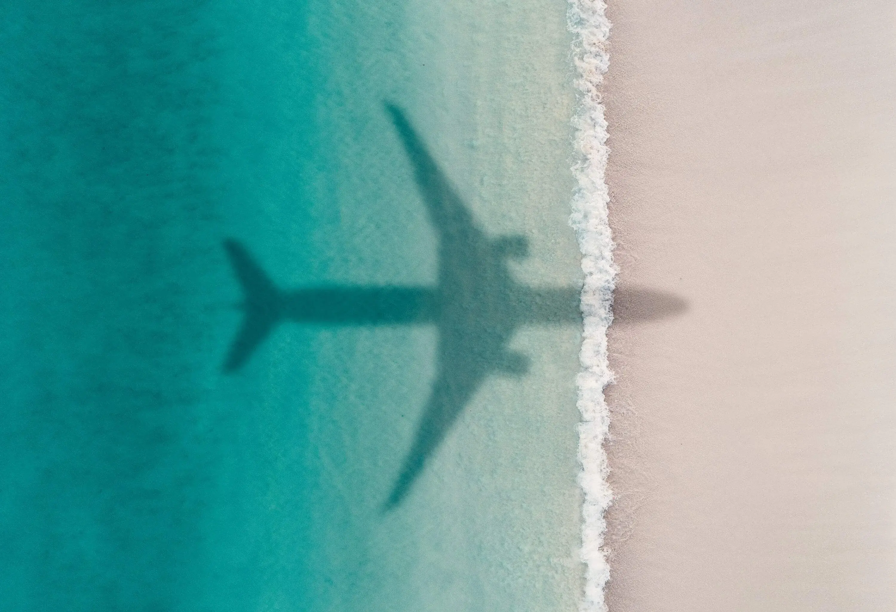 An airplane's shadow casts on a beach with turquoise waters.