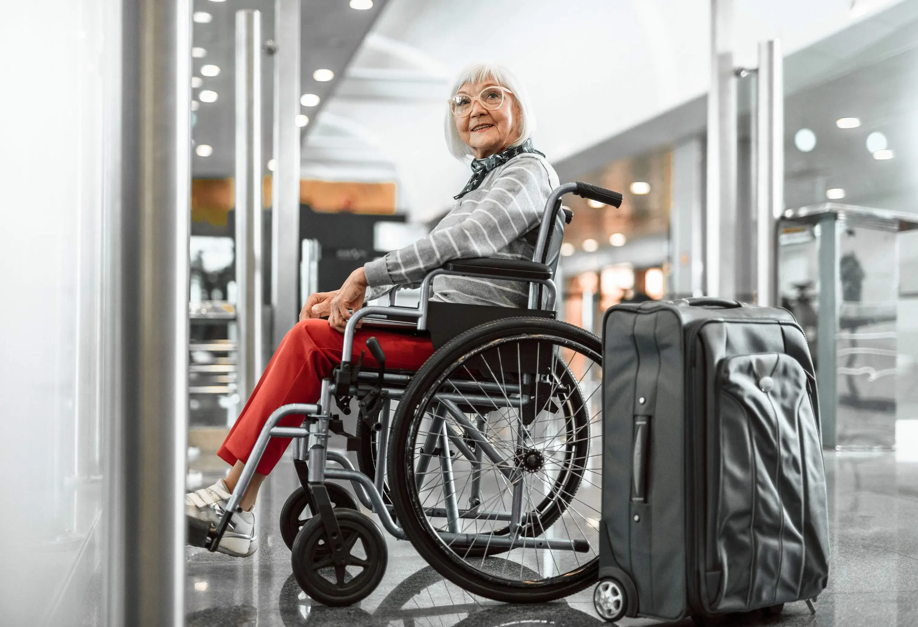 Full length portrait of mature female with laptop and suitcase sitting in wheelchair at airport and waiting for trip