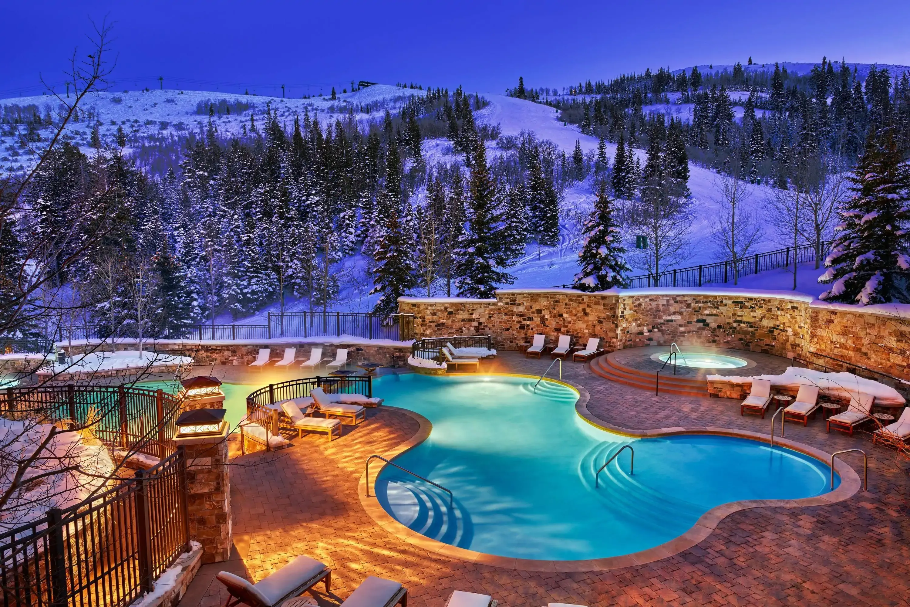 Night view of a lit outdoor hotel pool with snowy mountains and woods in the background