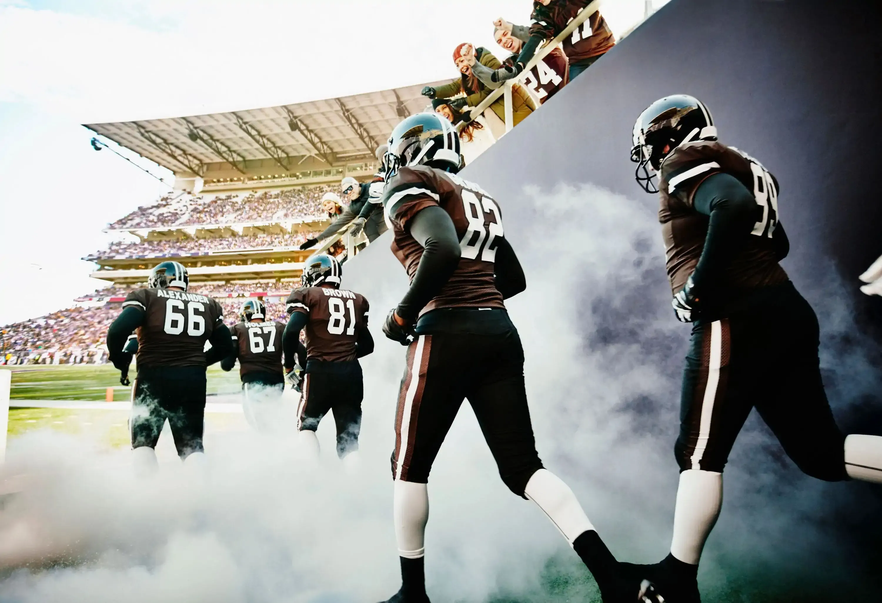 Fans cheering professional football team running out of tunnel through fog onto stadium field before game