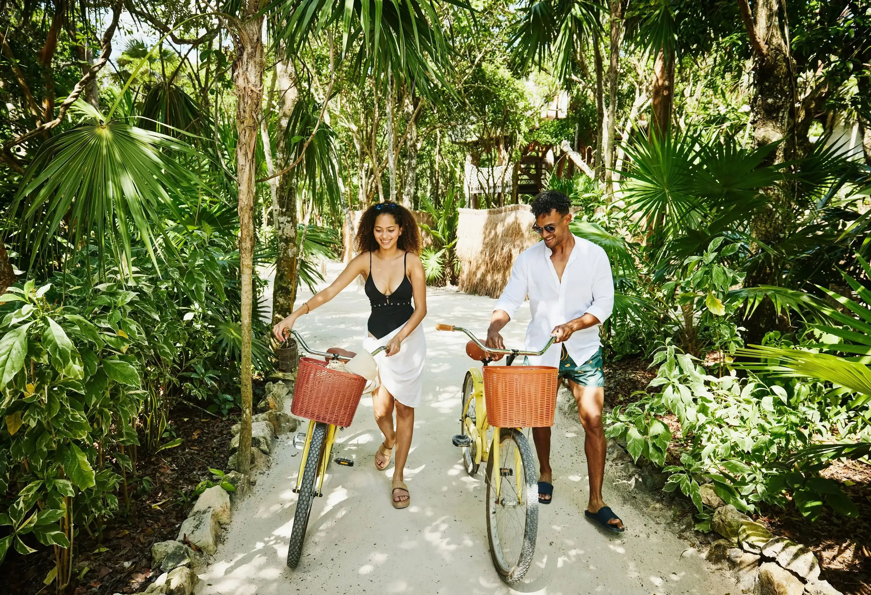 Two individuals leisurely lead their bicycles along a path enveloped by lush tropical plants.