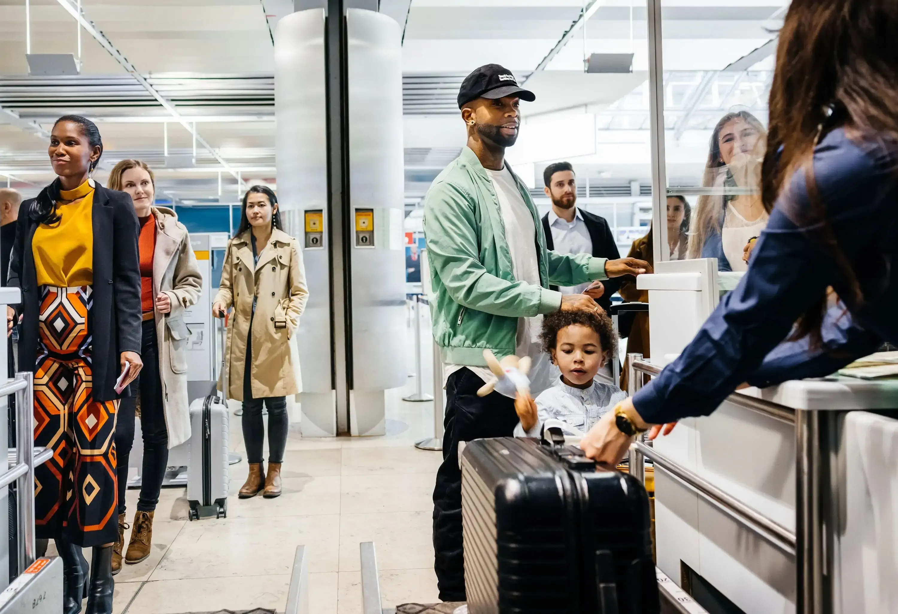 A family waits, among other travelers, in line at an airport luggage check-in desk with a father handing over documents while holding his child's hand.