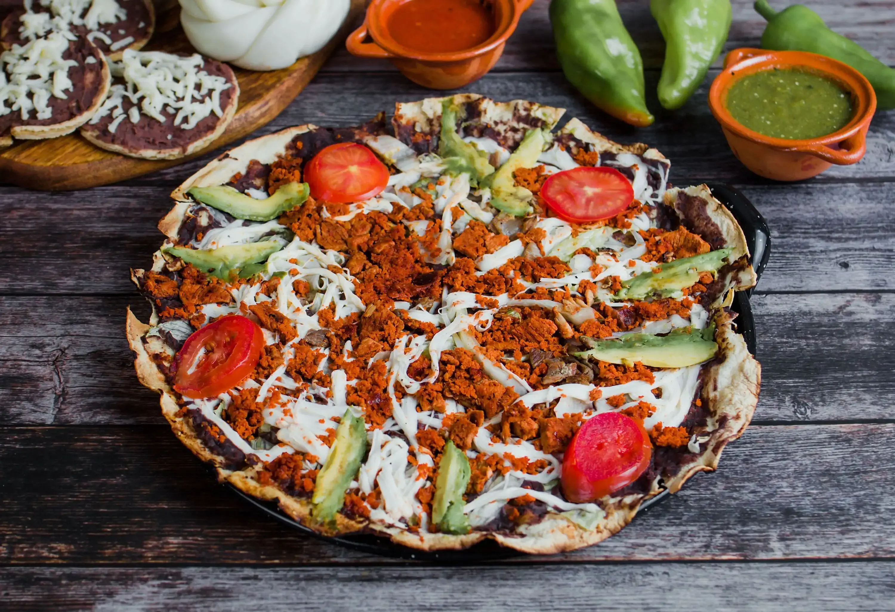 A traditional pizza-like dish toasted tortilla covered with tomato, avocado and cheese next to fresh chillies and dips.