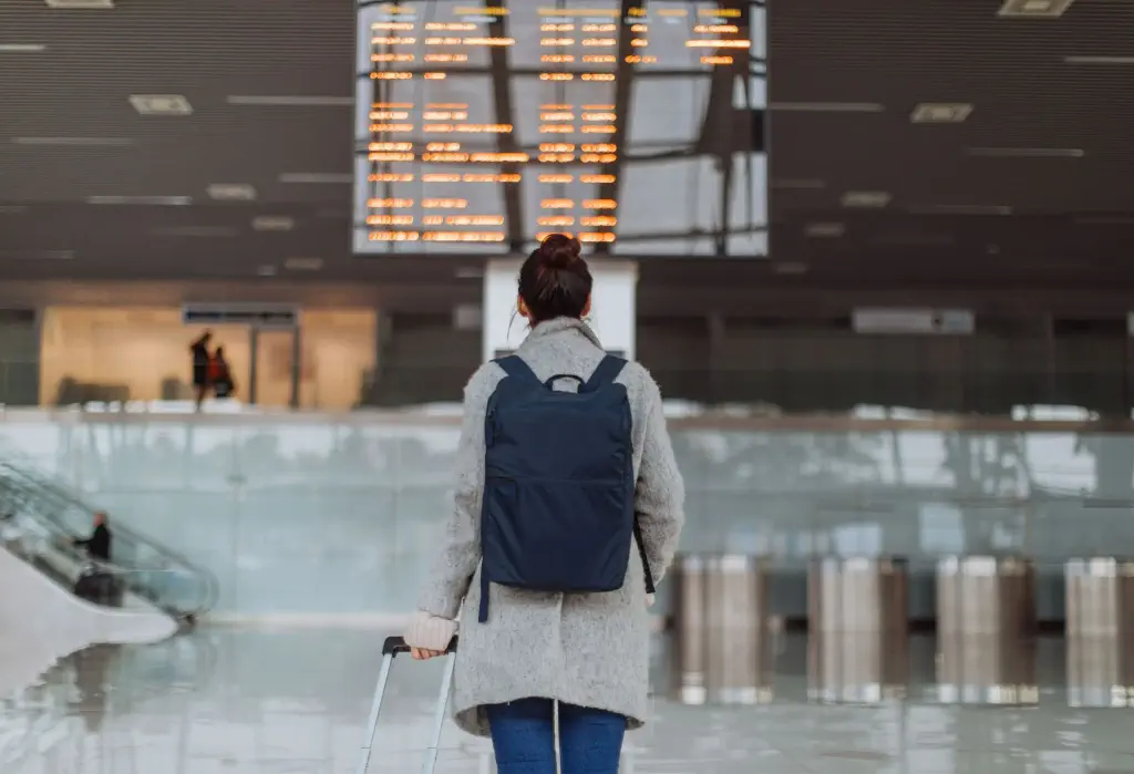 Woman at the airport, checking the arrival and departure boards for flight information while holding her luggage and carrying a backpack.