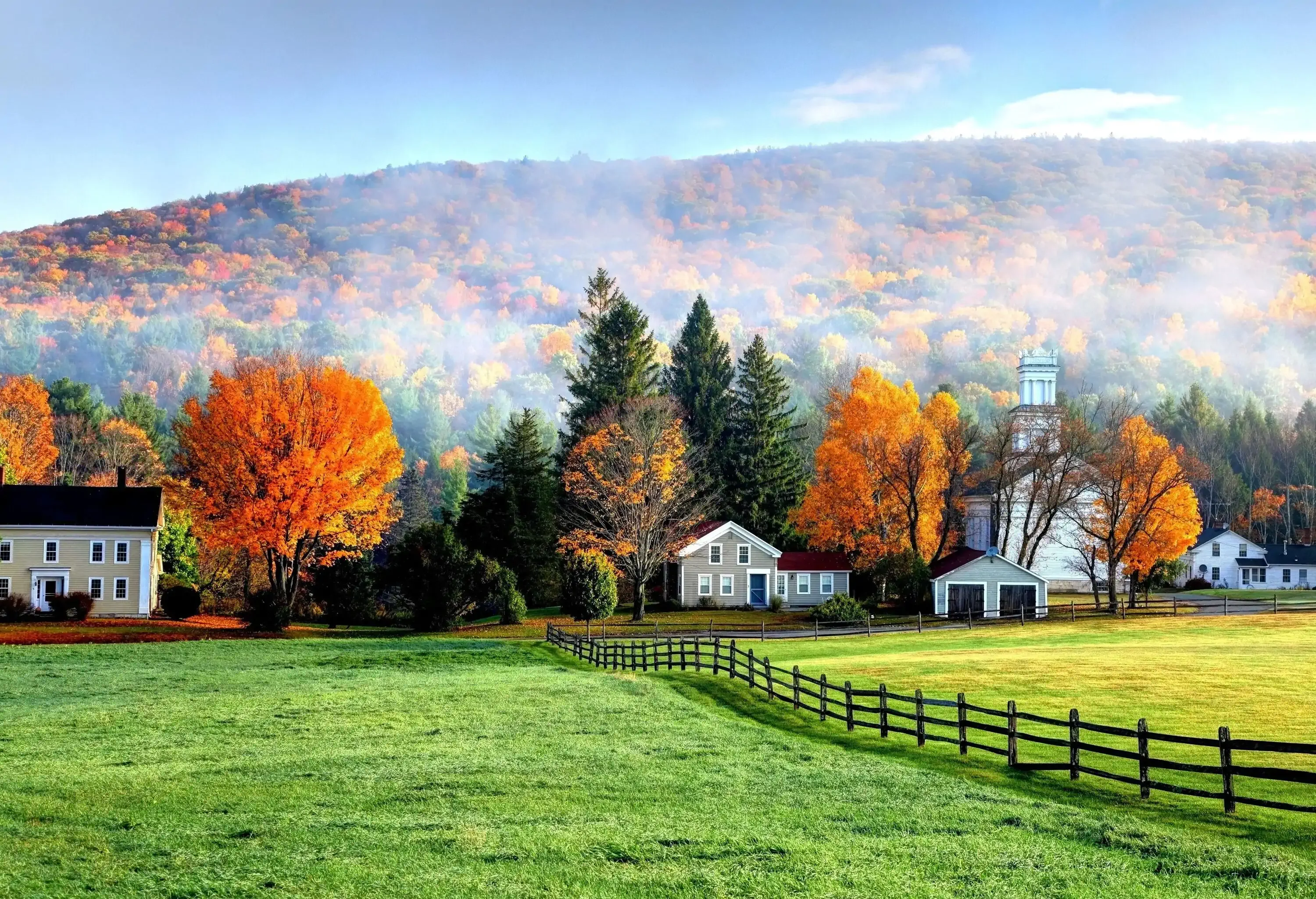 Green meadows with houses behind a forested hill boasting with autumn foliage.