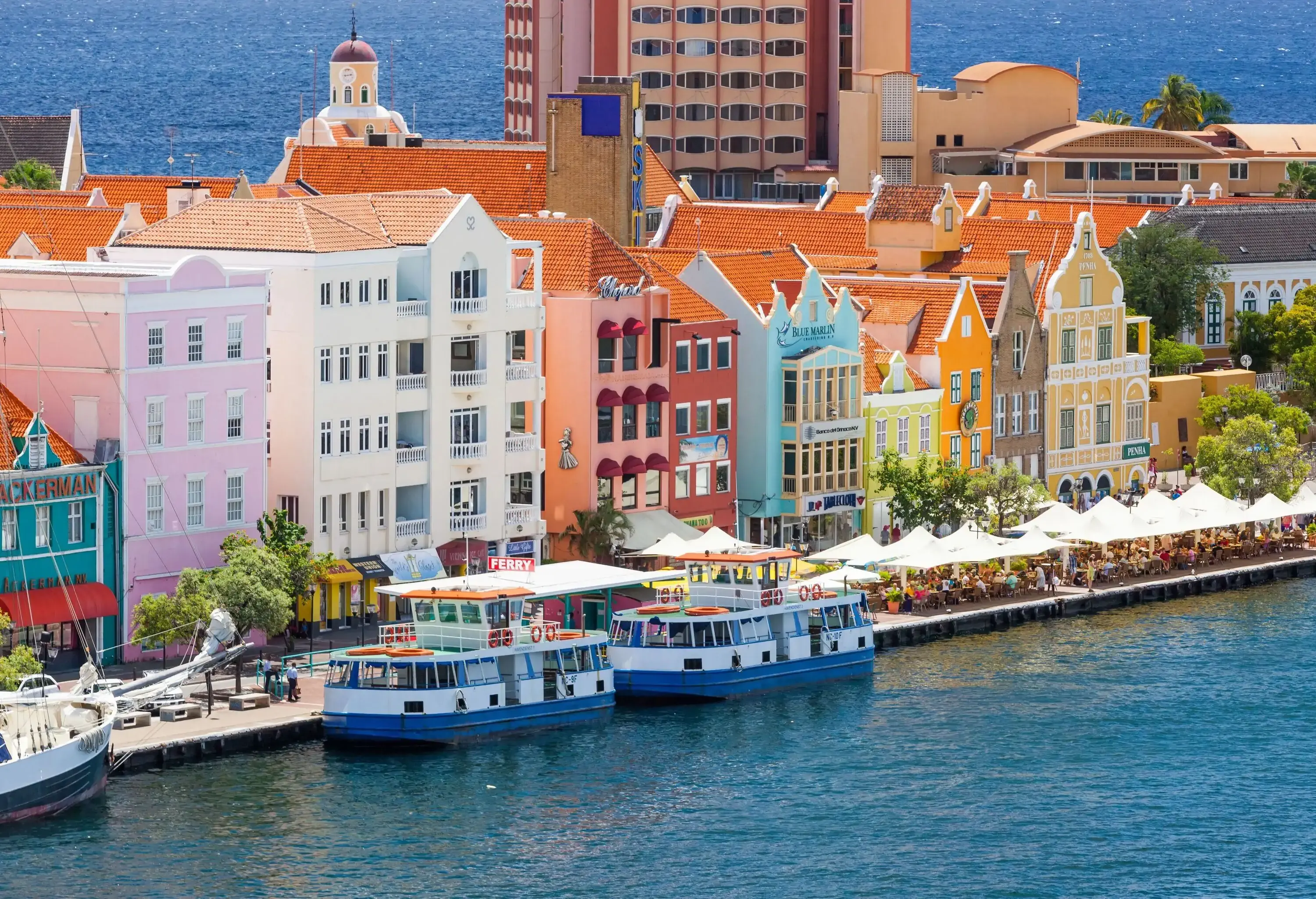 A picturesque line of waterfront buildings stands proudly along the shore, with ferries peacefully moored by the lively promenade, where a delightful line of outdoor dining establishments offers a perfect spot to savour the coastal ambiance.