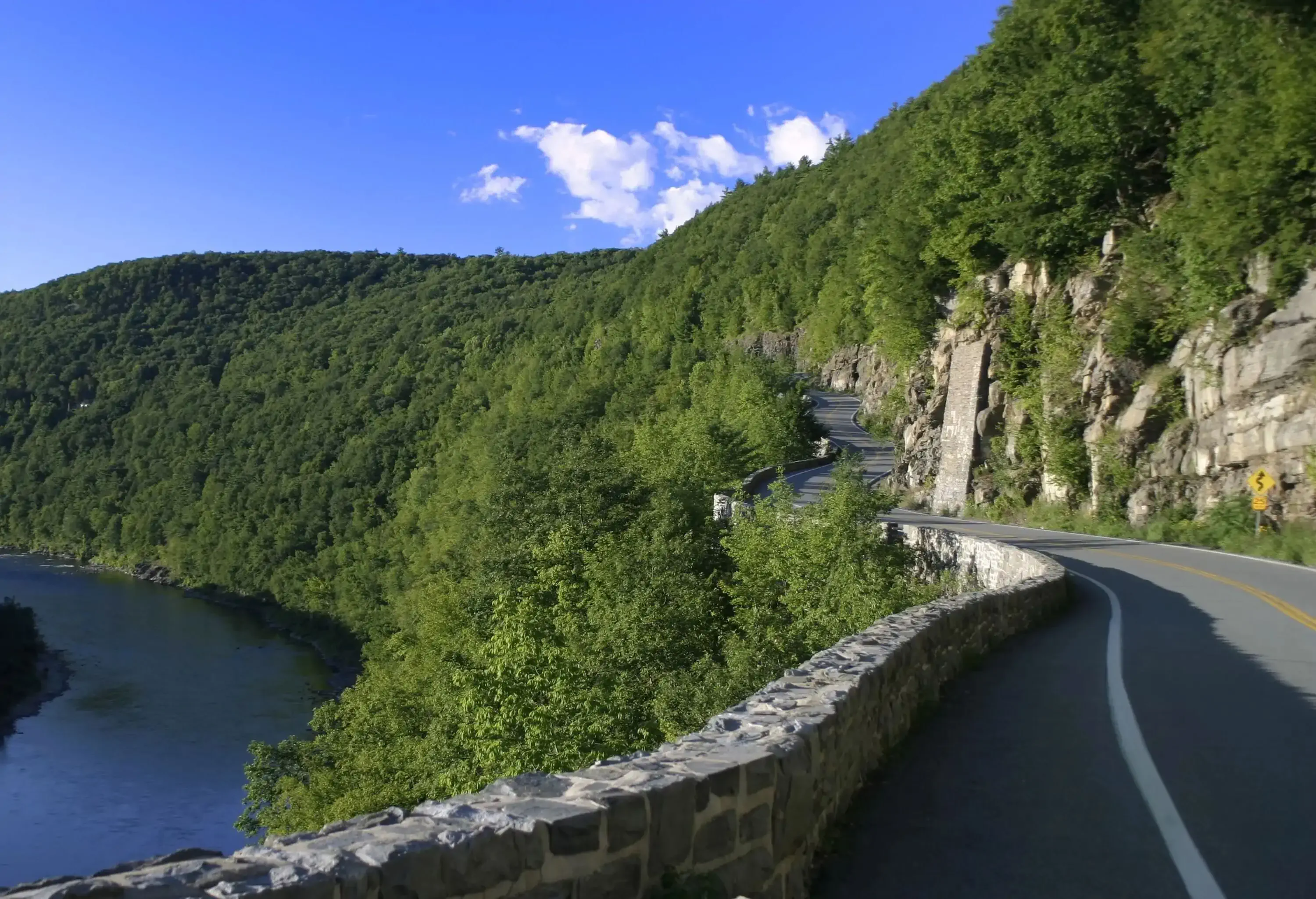 An empty road along a forested cliff with overlooking views of a calm river.