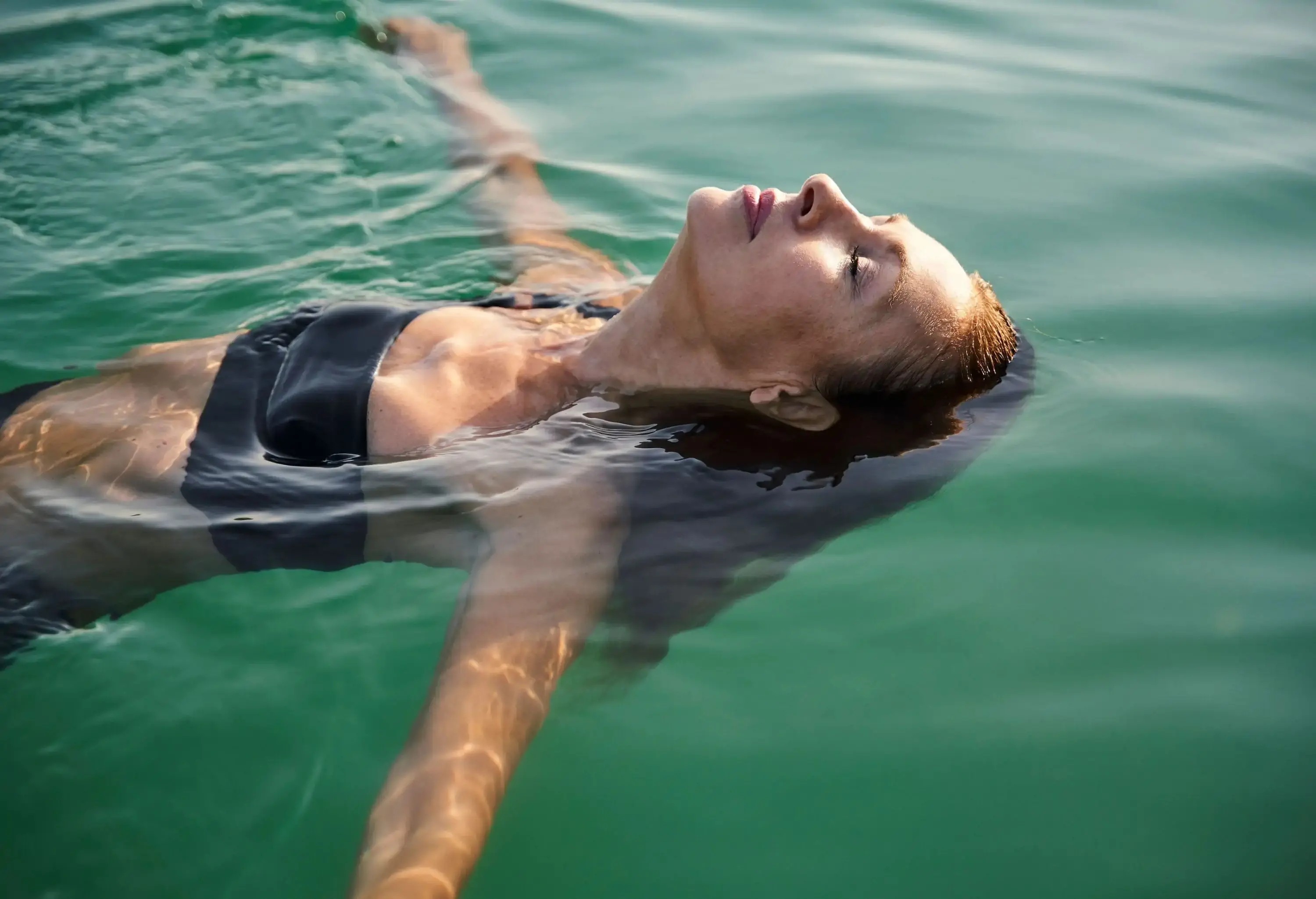 A brunette woman in black swimwear closes her eyes as she floats on the water.
