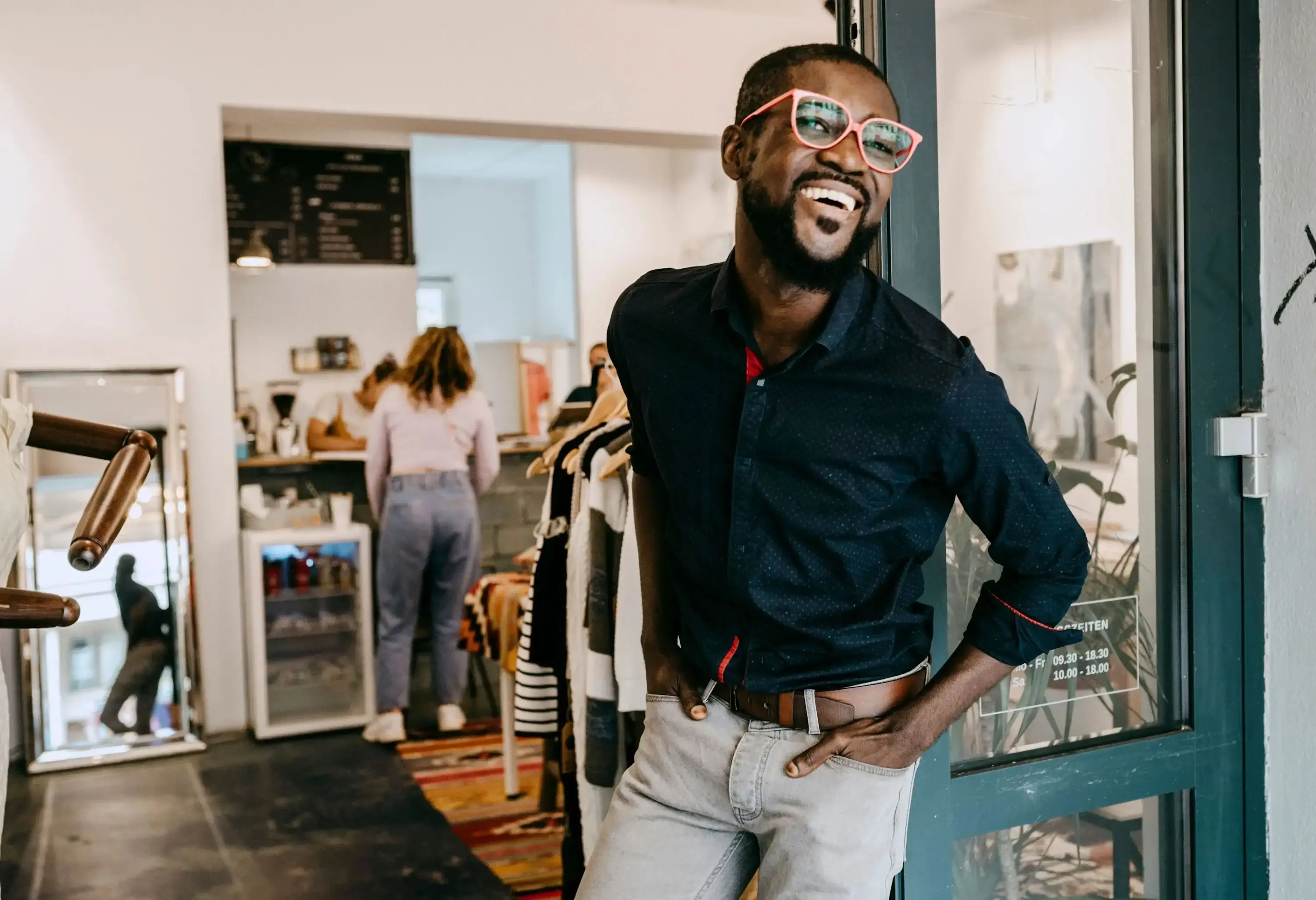 A friendly and enthusiastic store owner with his hands casually placed inside his pockets, standing at the entrance of a clothing store.