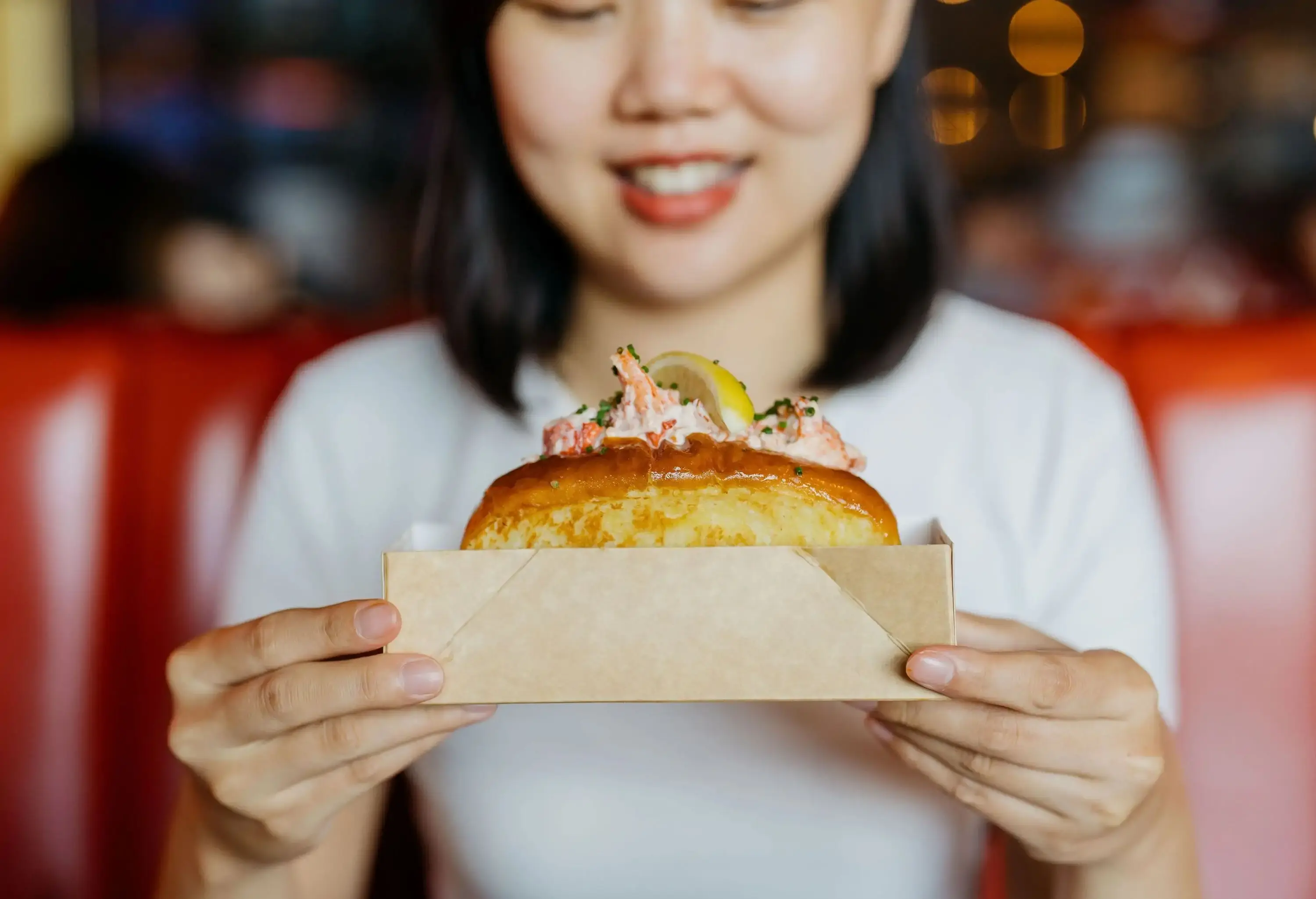 A woman smiles as she holds a lobster sandwich inside a paper box.