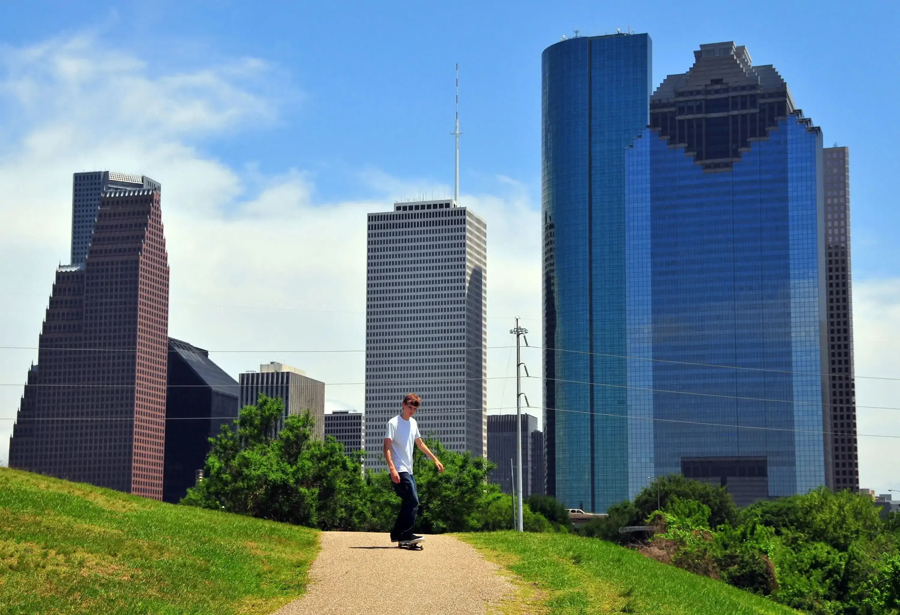 A young man skateboards on a hillside path against the backdrop of modern high-rise buildings.