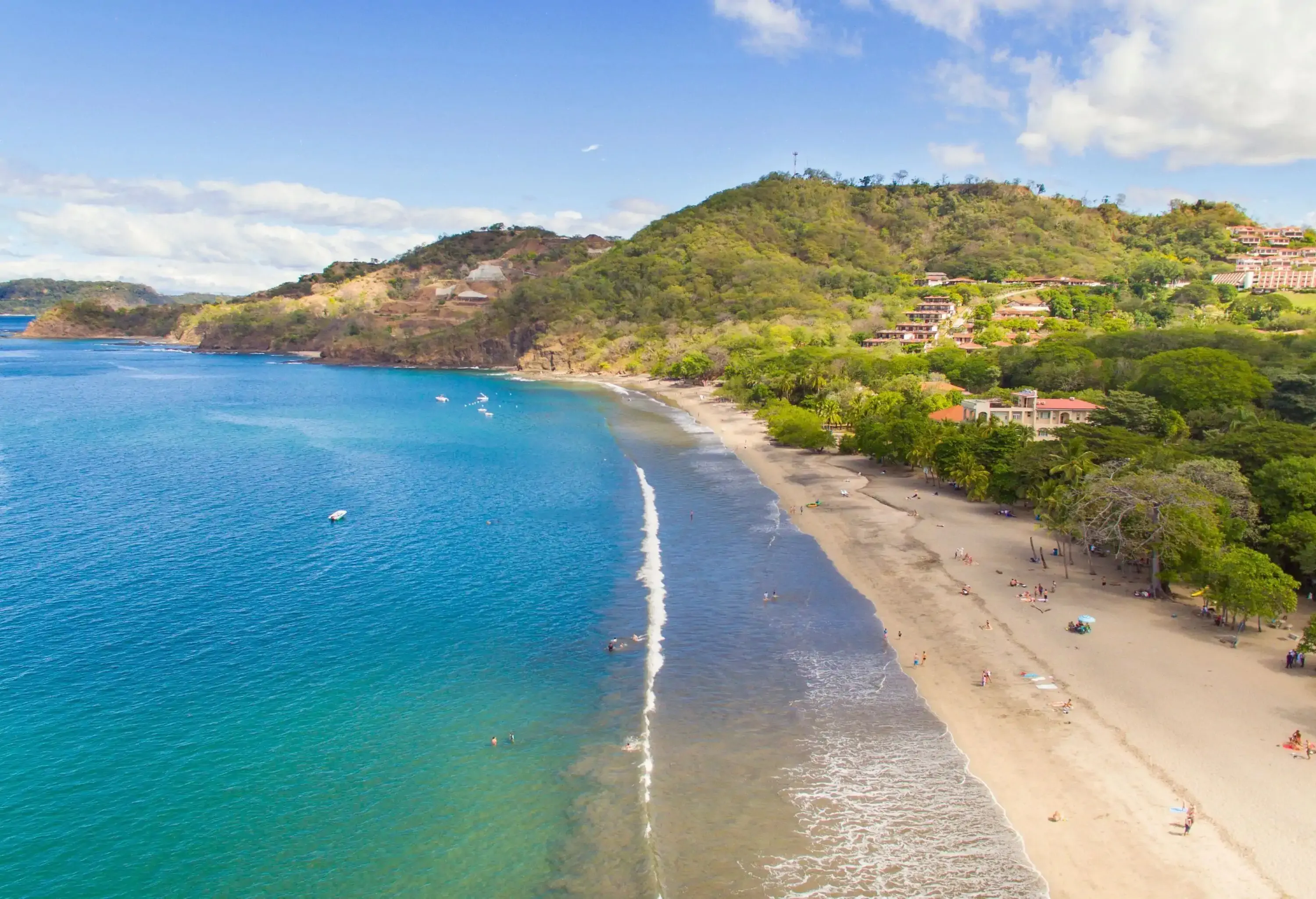 A breathtaking view of a turquoise and azure sea, embraced by a sandy shore dotted with beach goers, set amidst a forested island.