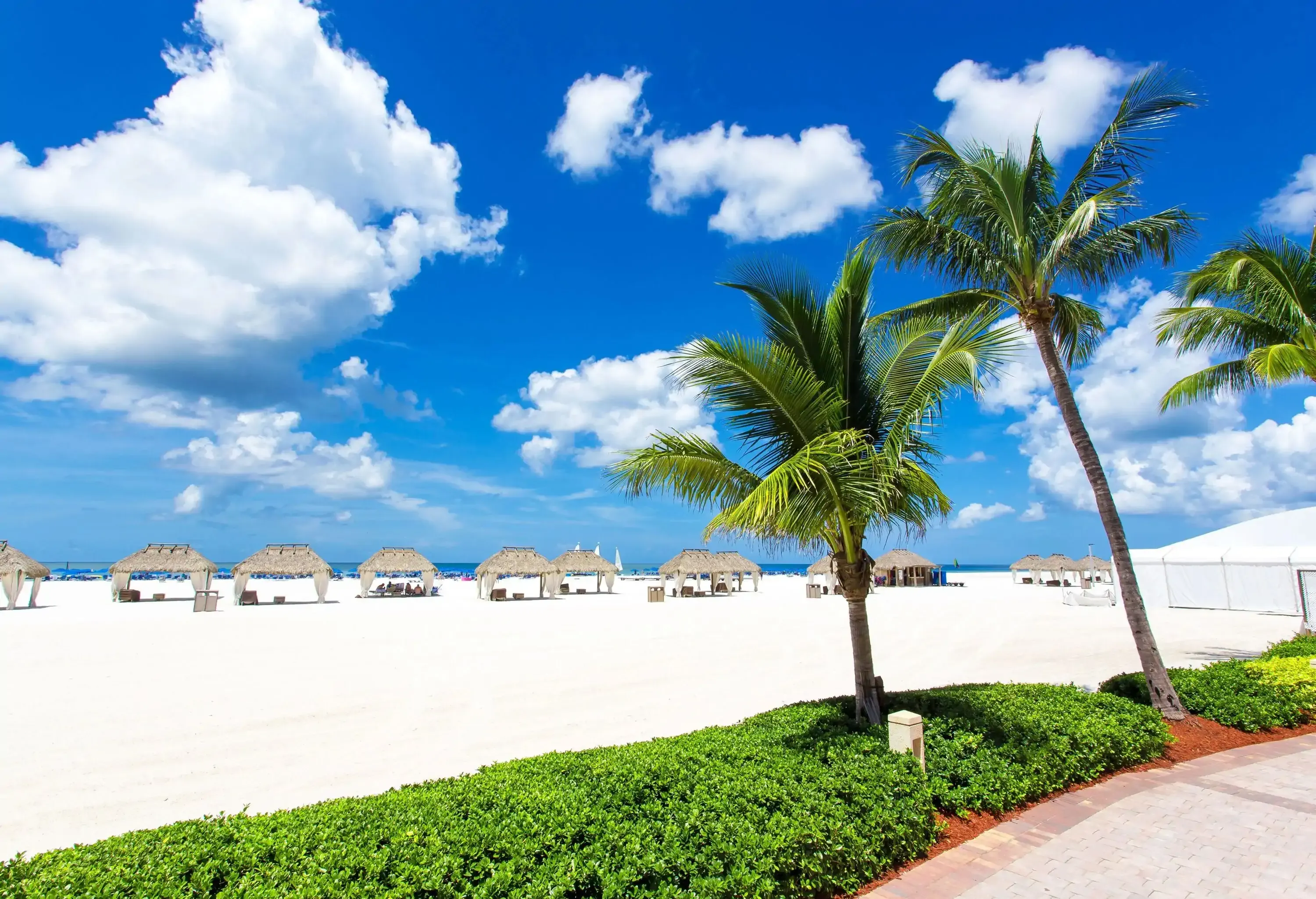 A white sand beach with hut umbrellas and sun beds against the cloudy blue sky.