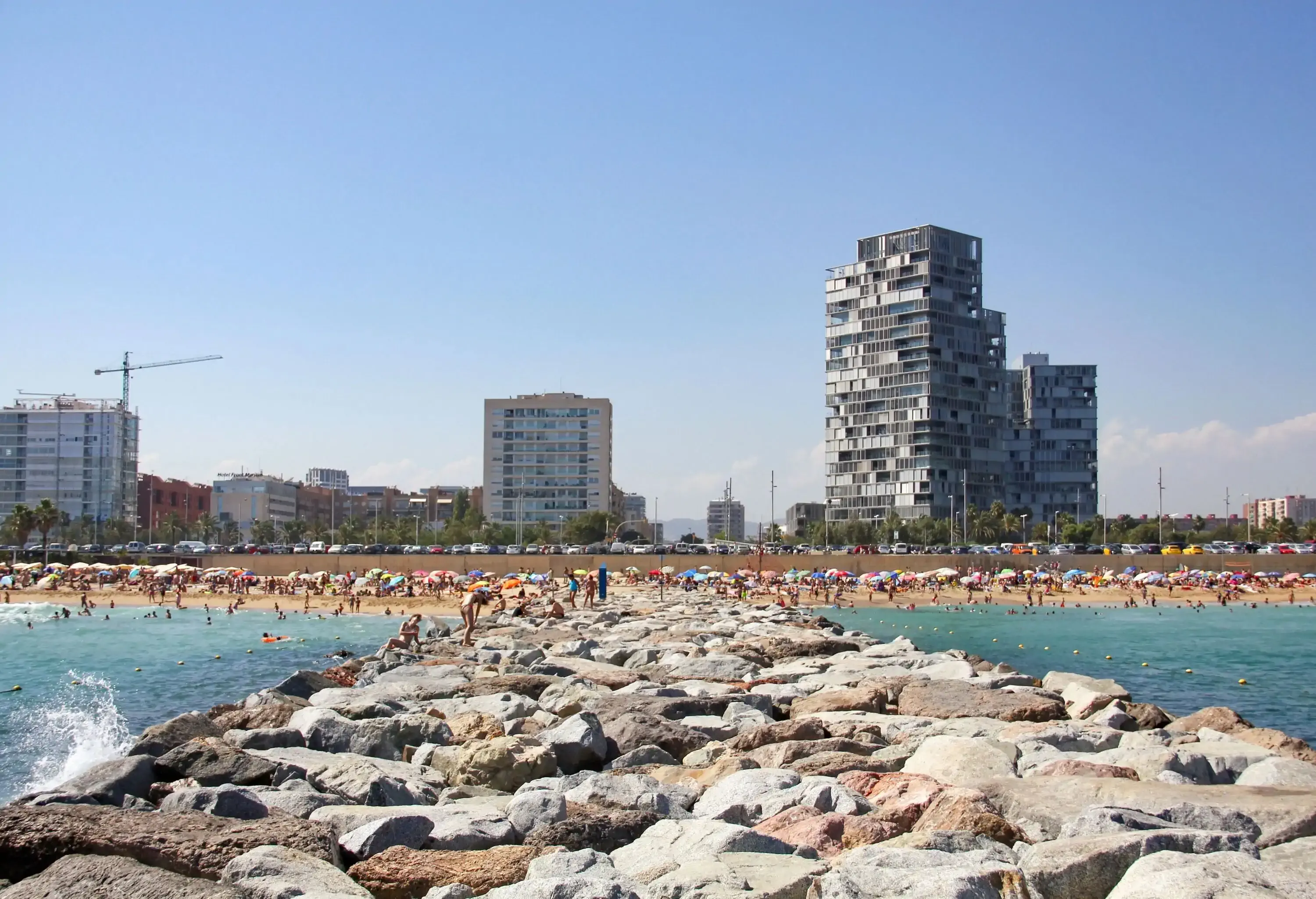 A crowded beach with a broad stone breakwater across the sea.