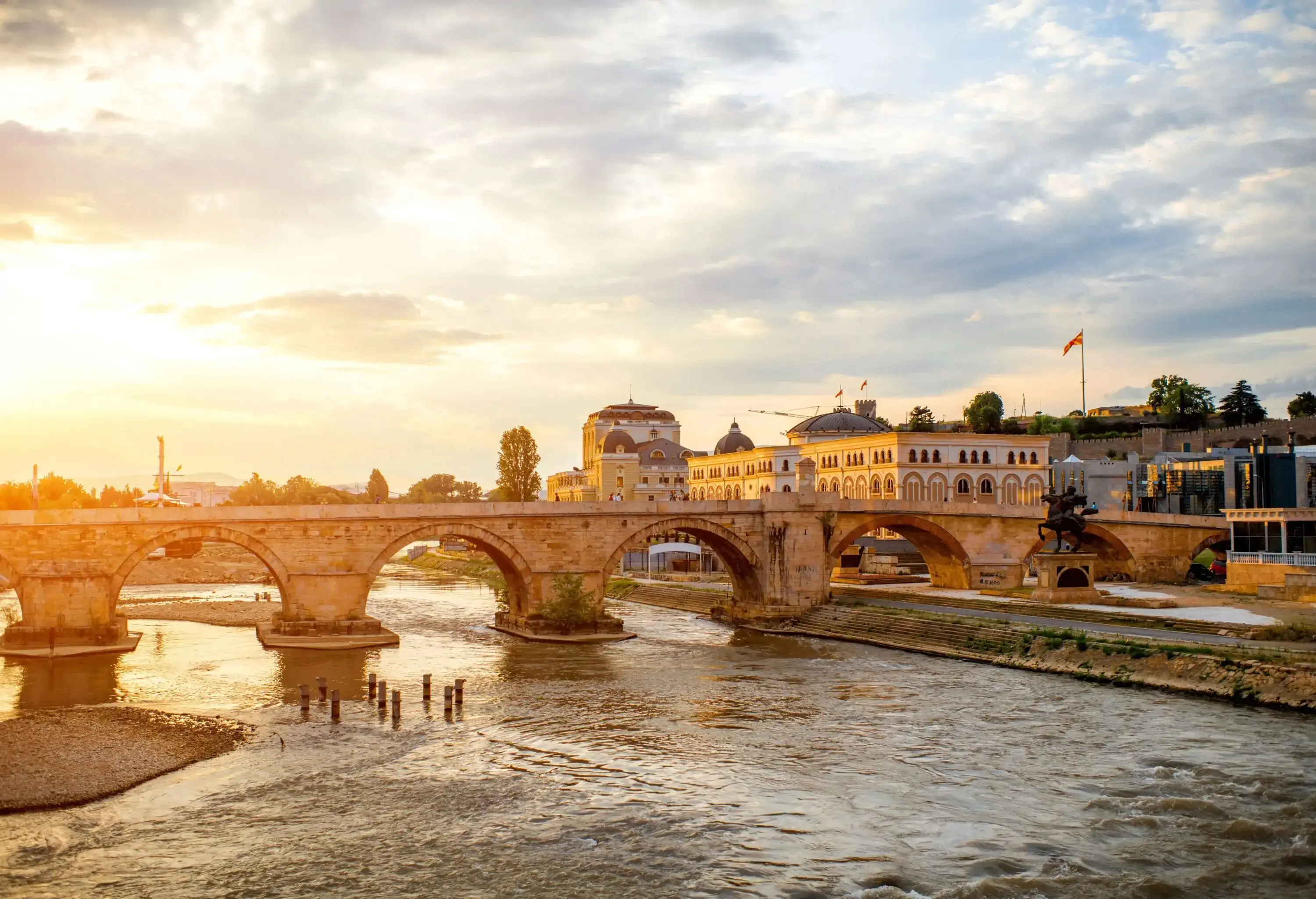 The scenic sun shines through a stone arch bridge that spans the river along the old buildings.