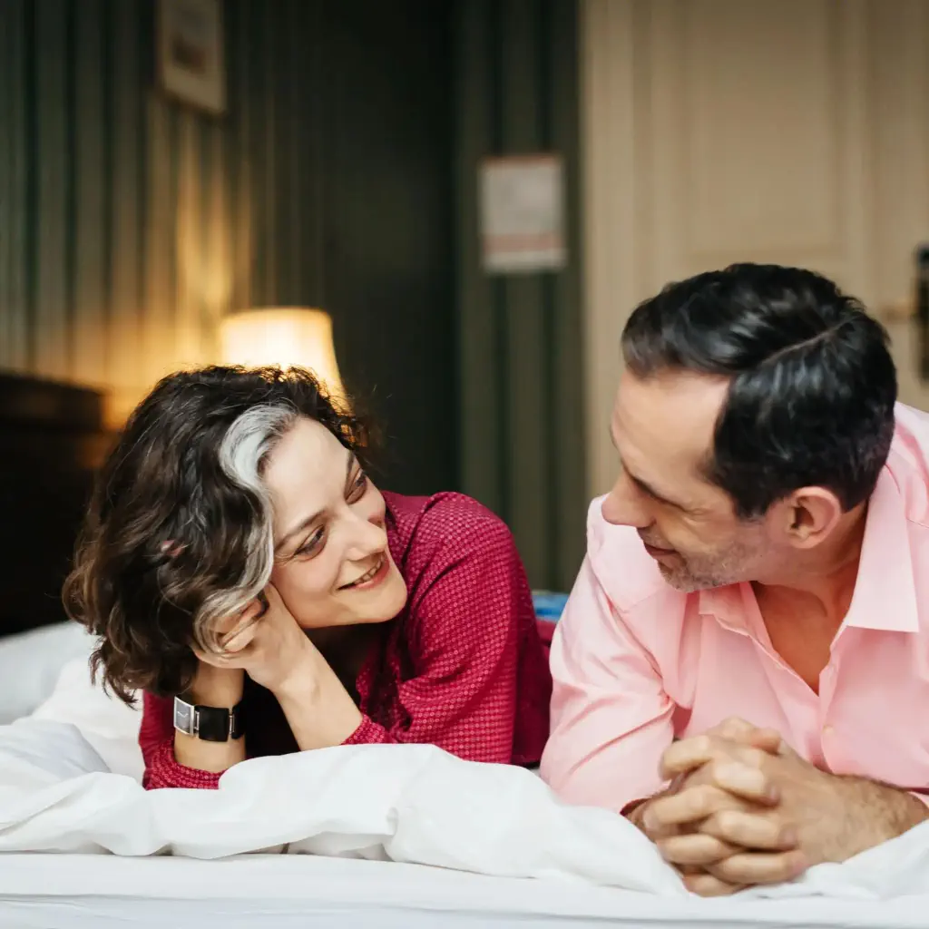 A mature, happy couple is lying at a hotel bed next to each other. They both are smiling and seem to be in love. In the background, the dark and green interior of the room can be seen.