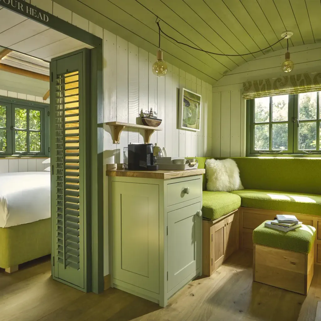 Cozy wooden cabin interior with green and white decor. Features a bed, corner sofa with fluffy pillows, small kitchen area, and bright windows. Warm, inviting ambiance.