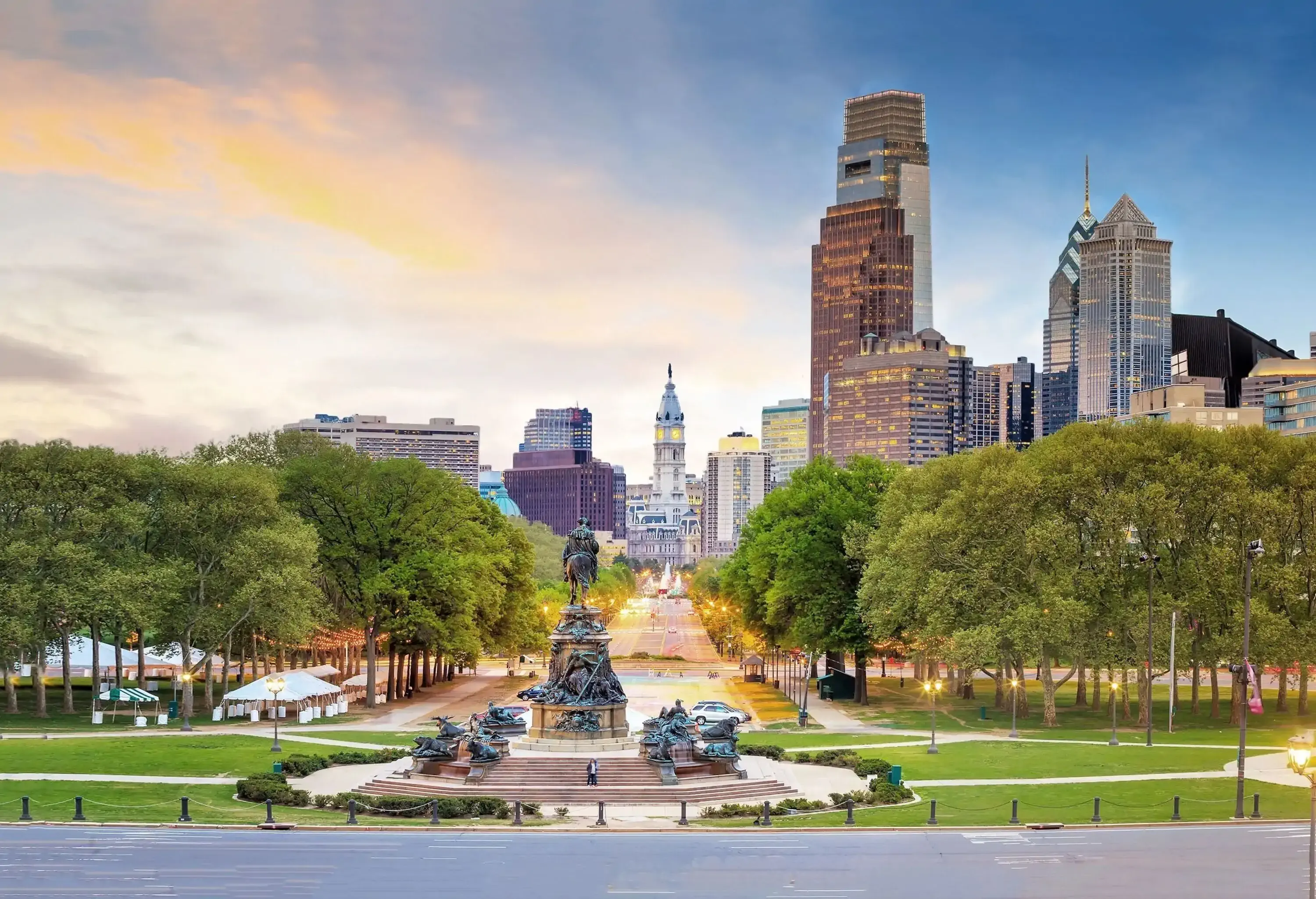 A massive statue in the middle of a park with lush trees and views of the city skyline under the sunset skies.