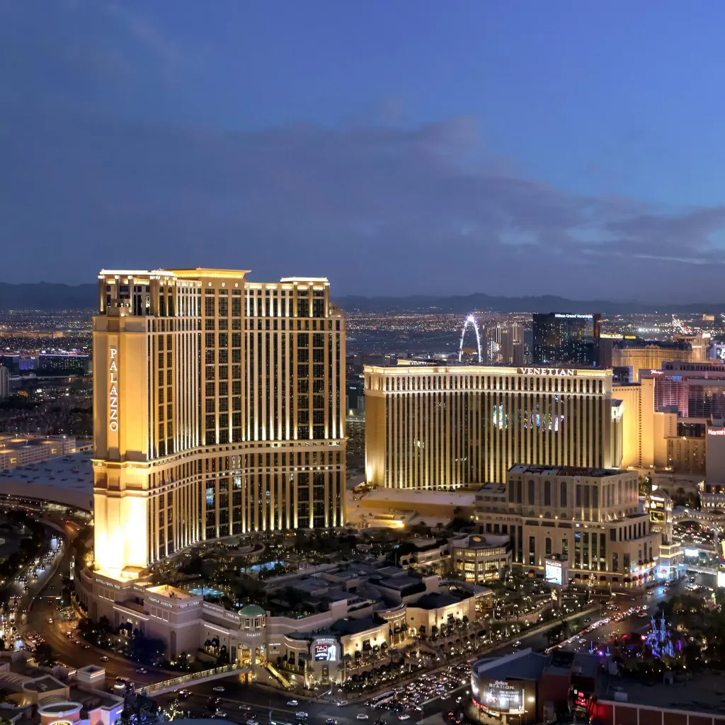 Aerial view of Las Vegas at dusk, featuring the illuminated Palazzo and Venetian hotels. The city skyline glows warmly under a darkening blue sky.