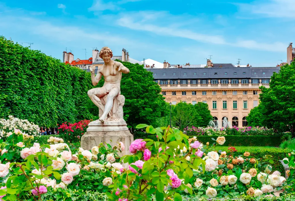 Marble statue in a rose-filled garden at Palais-Royal, Paris, with classic building in background.