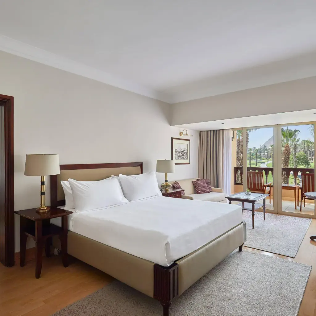 Hotel room with a large bed, wooden furniture, and balcony overlooking palm trees and gardens. Soft lighting creates a calm, inviting atmosphere.