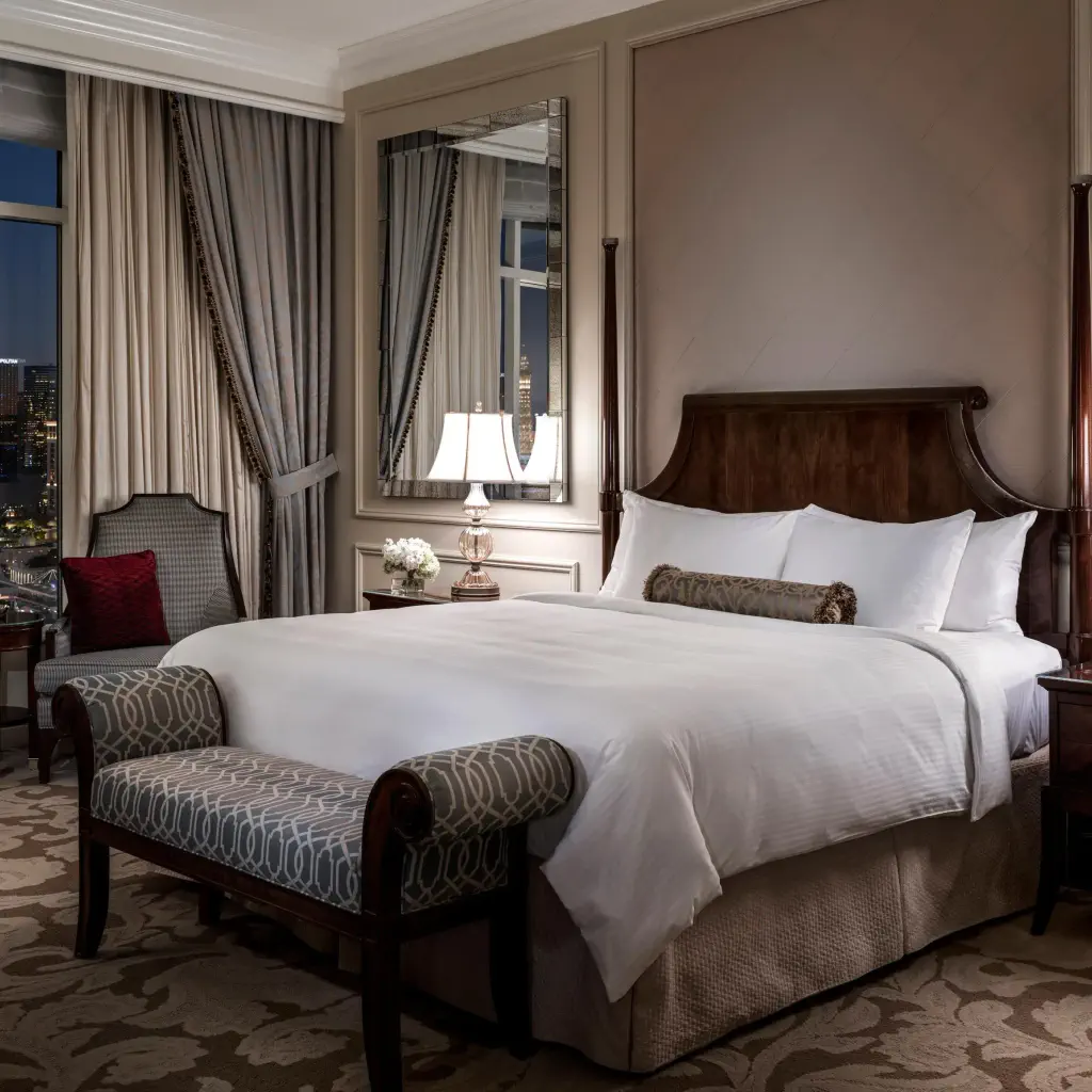 Elegant hotel room with a large, plush bed and ornate wooden headboard. Two lamps on end tables, bench at the foot, and floor-to-ceiling window with city view. Luxurious ambiance.